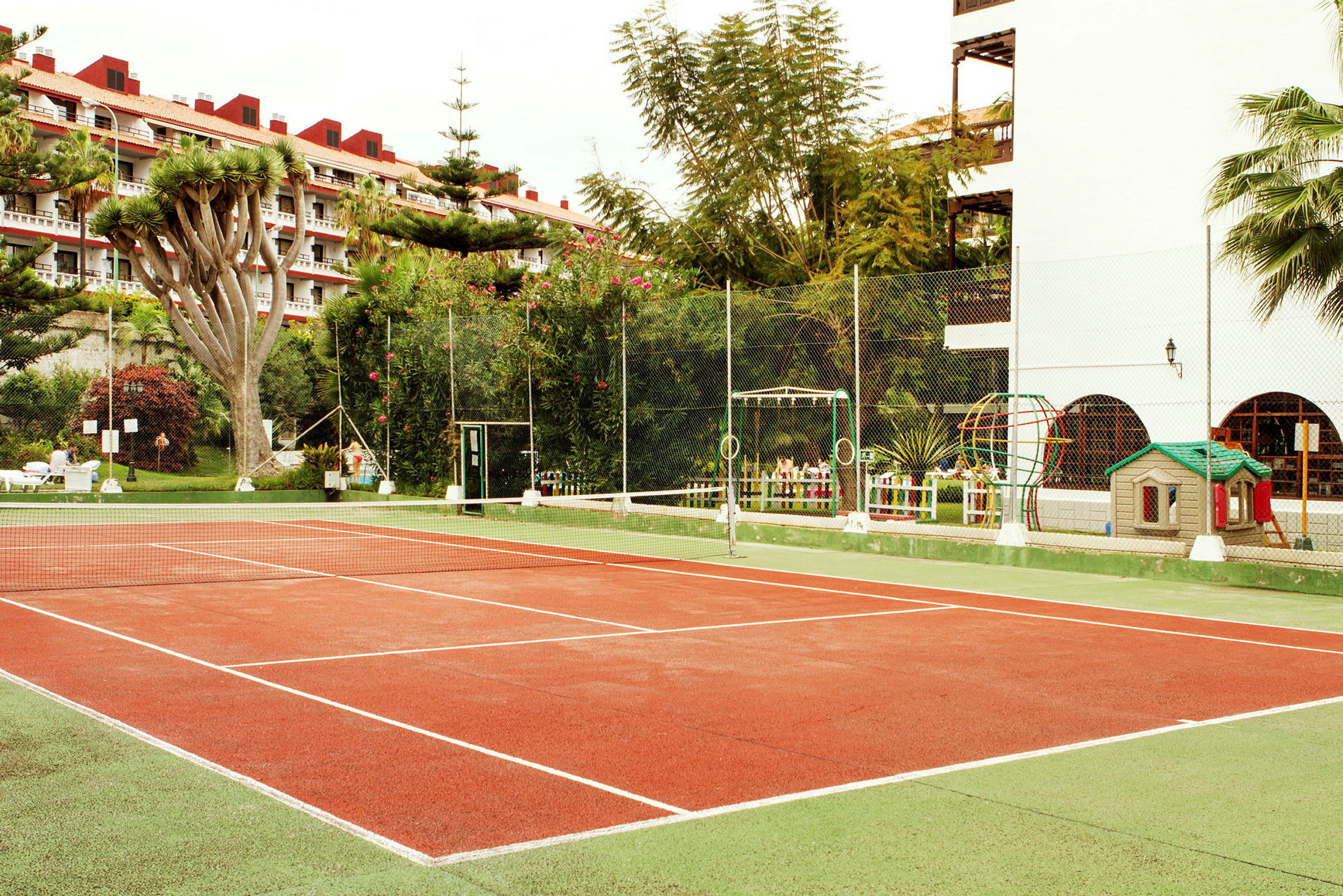 Tenniskenttä, jota ympäröivät kasvillisuus ja rakennukset. (Tekoälyn luoma teksti)