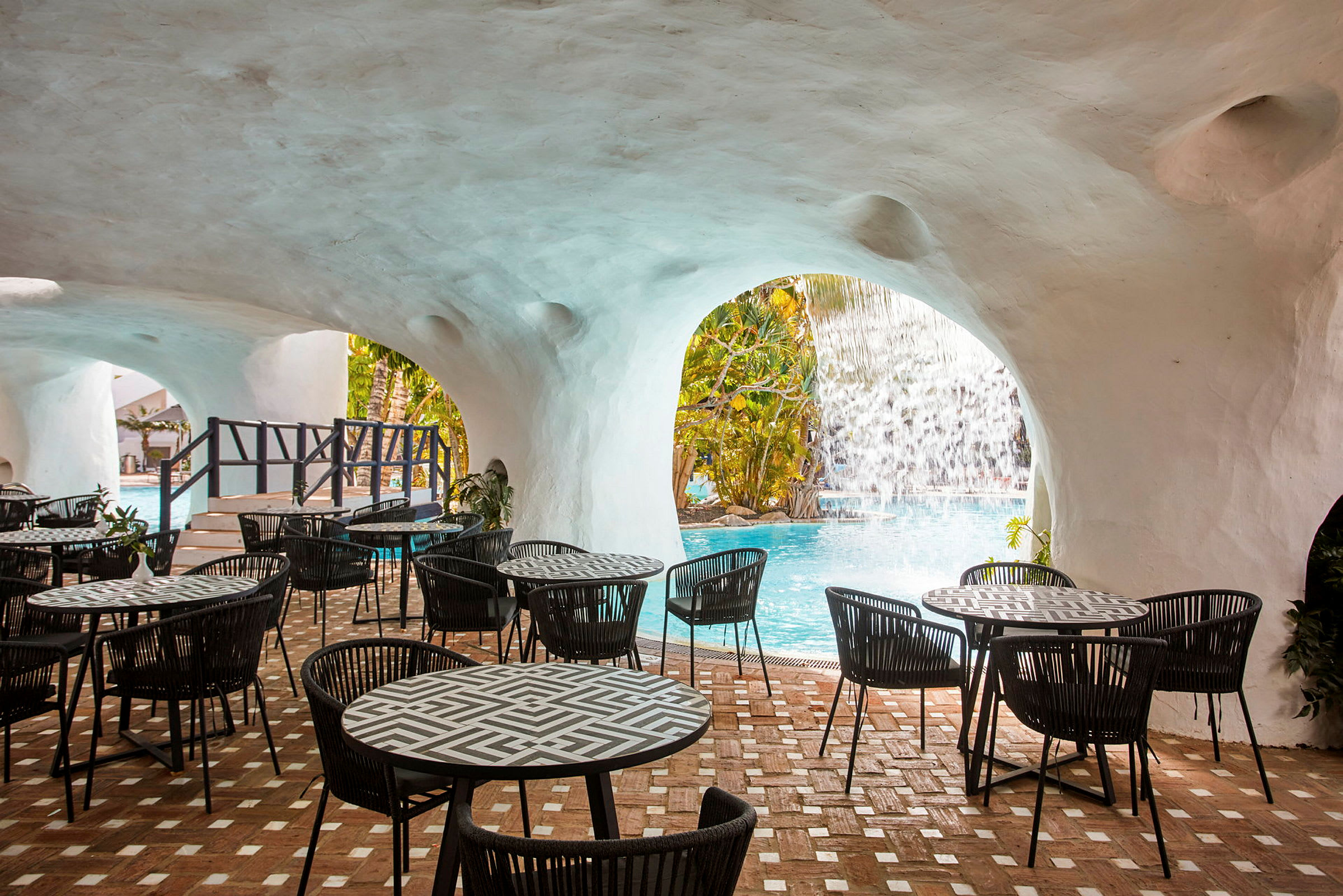 Ulkoravintola, jossa pöytiä ja tuoleja kaarevan katon alla, näkymä vesiputoukselle altaan äärellä. (Tekoälyn luoma teksti)
