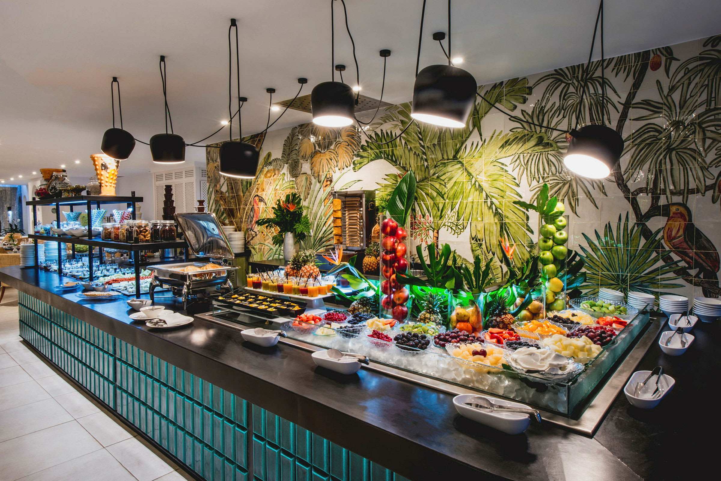 Buffet-pöytä, jossa on erilaisia hedelmiä, jälkiruokia ja koristeita modernin valaistuksen alla. (Tekoälyn luoma teksti)