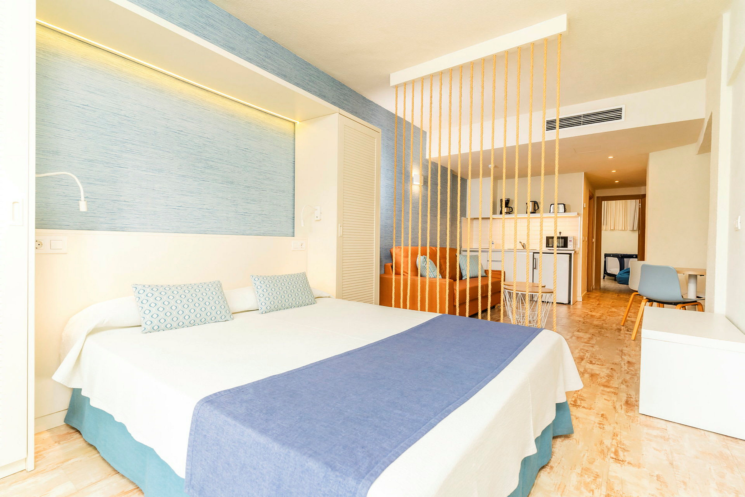 Hotellihuone, jossa on parivuode, sininen päiväpeite ja koristeelliset tyynyt, sekä oranssi sohva koristeellisen väliseinän takana. (Tekoälyn luoma teksti)