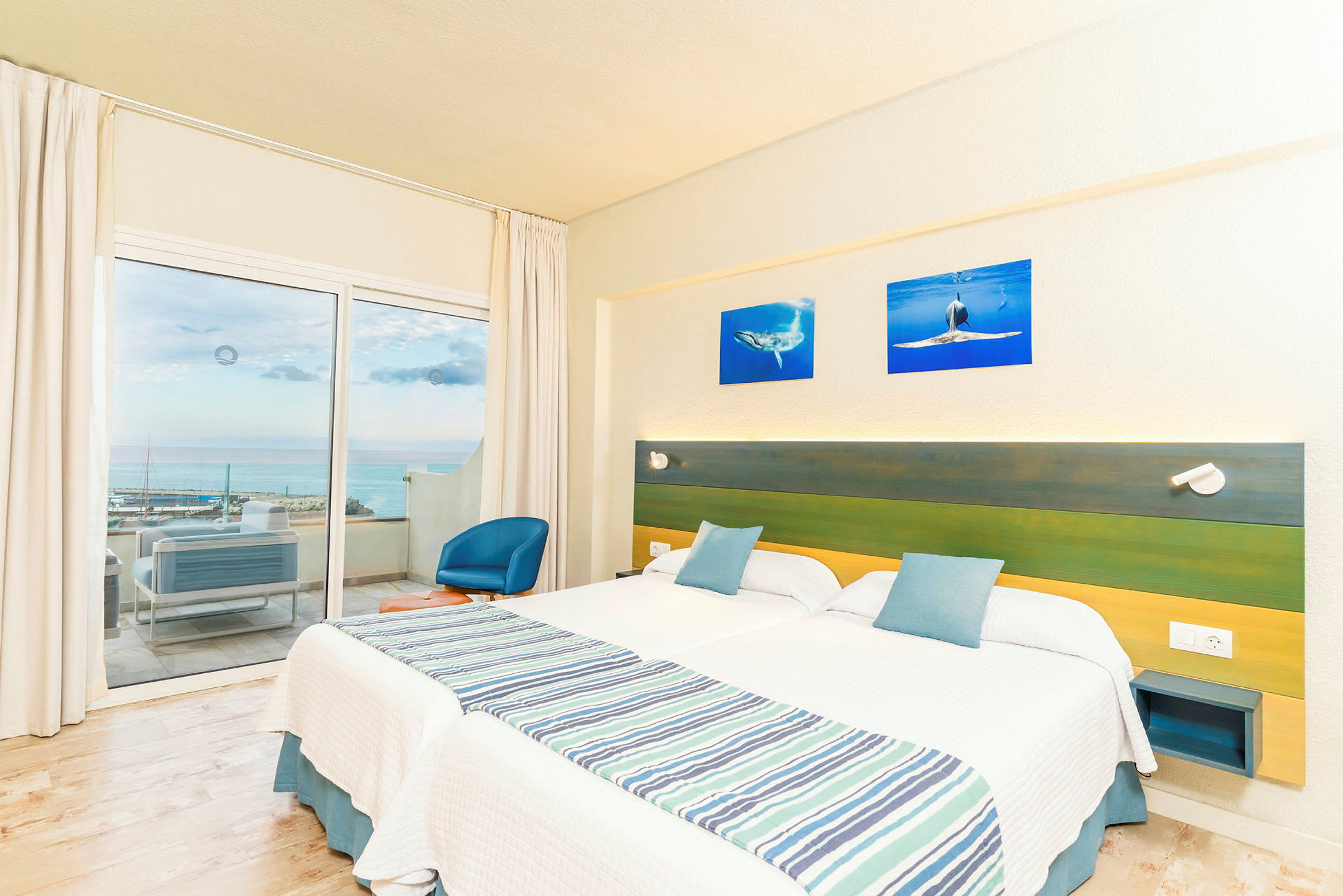Hotellihuone, jossa kaksi erillistä sänkyä, vihreä ja sininen sängynpääty sekä parveke merinäköalalla. (Tekoälyn luoma teksti)
