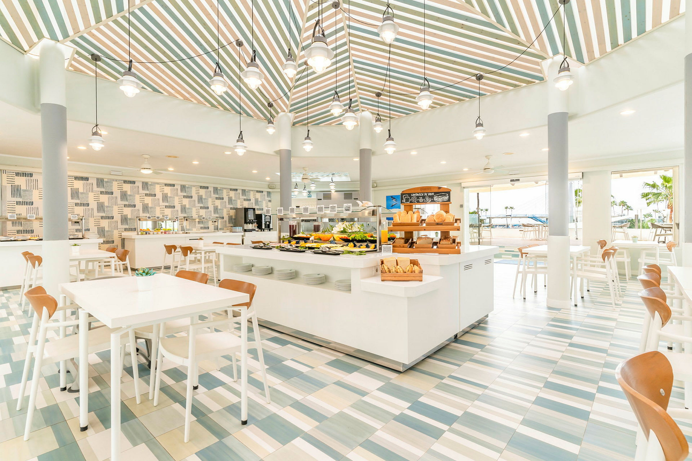 Valoisa ja moderni ravintola, jossa on valkoiset kalusteet ja värikkäät kaakelilattiat. Keskellä sijaitsee seisova pöytä erilaisilla ruoilla. (Tekoälyn luoma teksti)