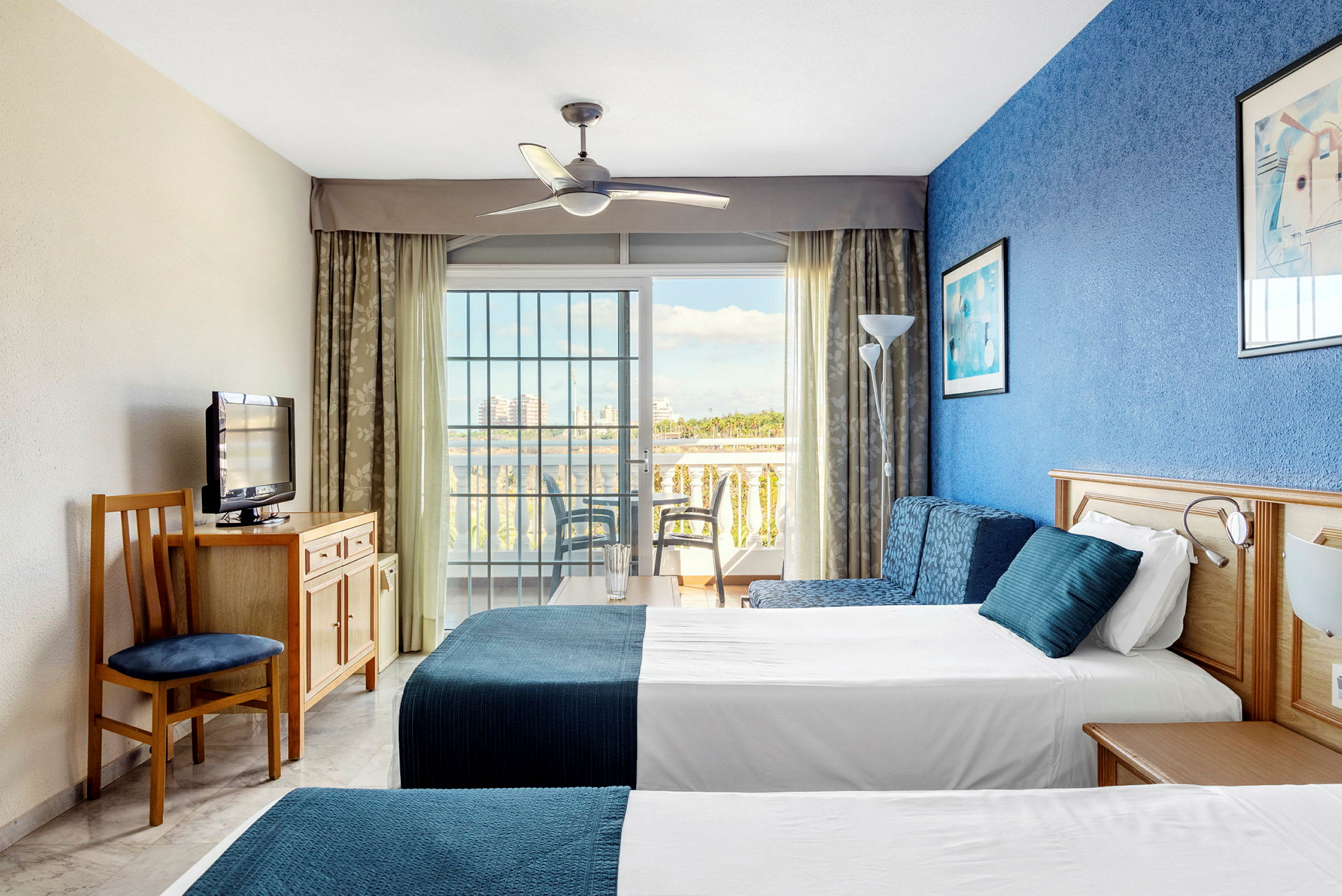 Hotellihuoneen kuva, jossa on kaksi vuodetta, sininen seinä ja lasiovi parvekkeelle. (Tekoälyn luoma teksti)