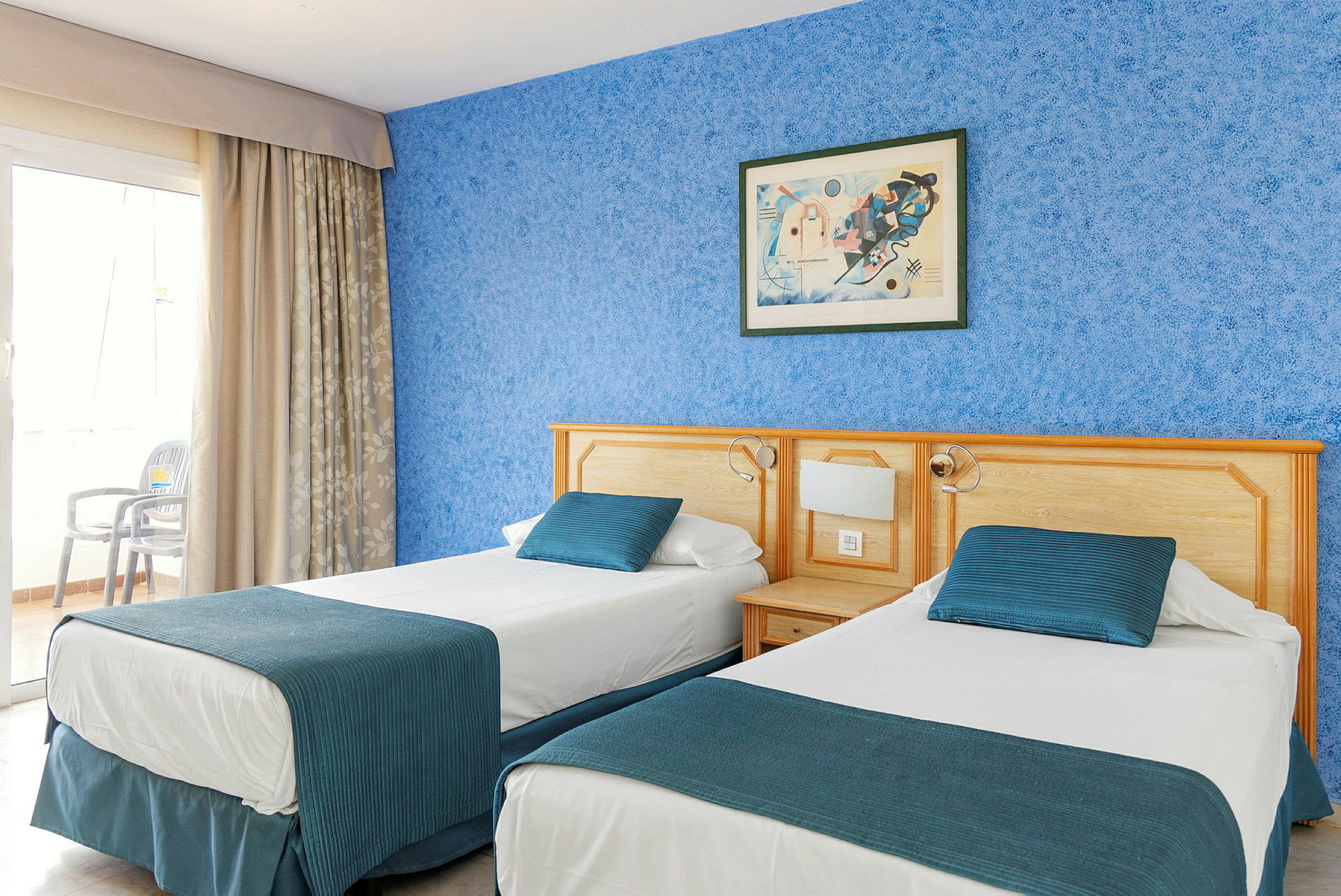 Hotellihuone, jossa on kaksi yhden hengen vuodetta, sininen seinä ja maalaus sängyn yläpuolella. (Tekoälyn luoma teksti)