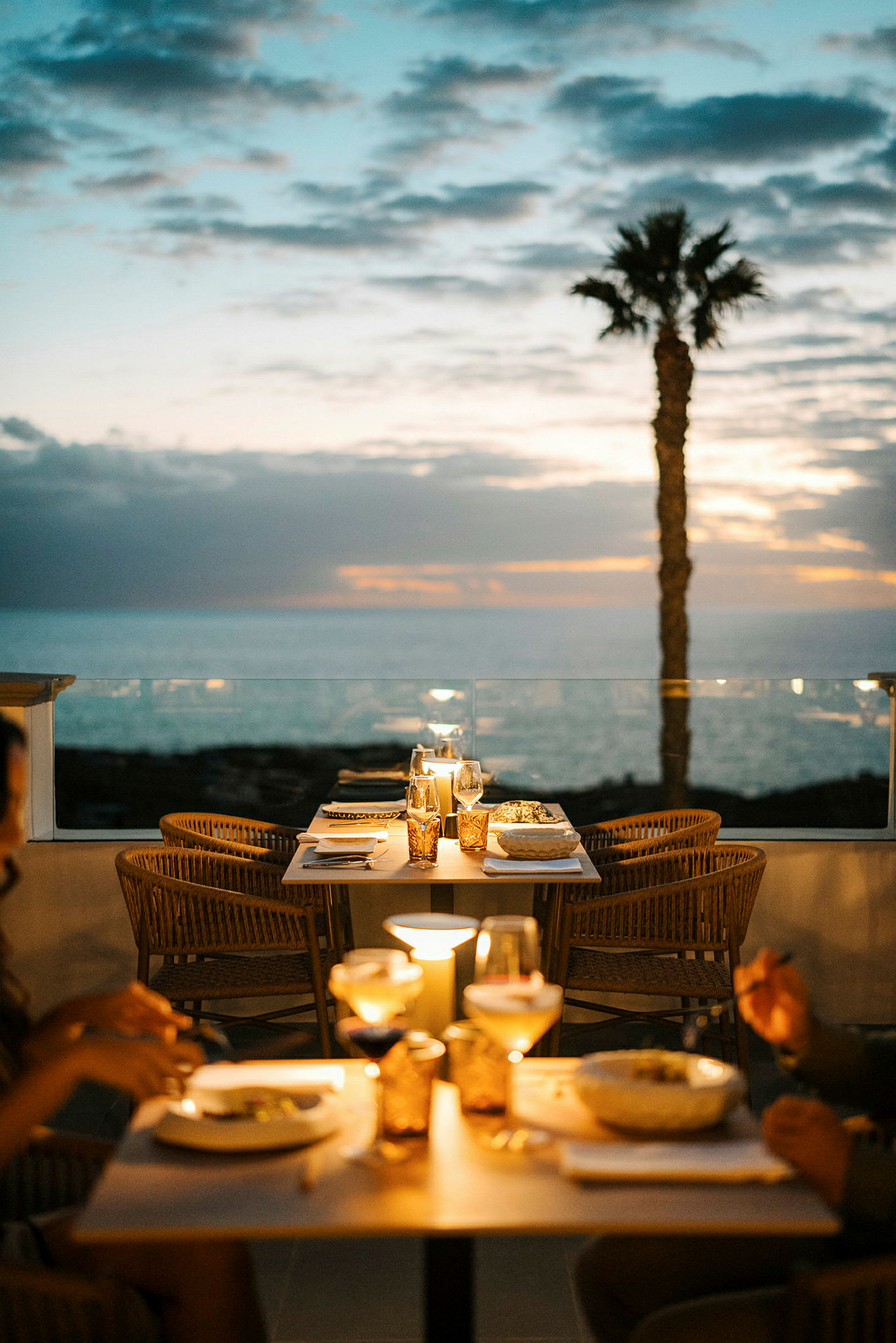 Ulkoterassi, jossa on pöytiä ja tuoleja, sijaitsee meren äärellä iltataivaan alla palmun ollessa taustalla. (Tekoälyn luoma teksti)