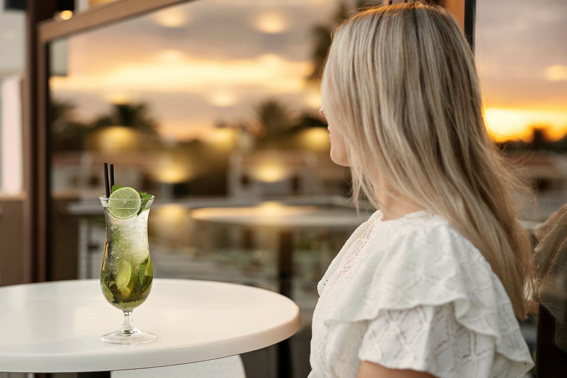 Nainen, jolla on pitkä vaalea tukka, istuu pöydän ääressä, jossa on cocktail edessä, taustalla auringonlasku. (Tekoälyn luoma teksti)