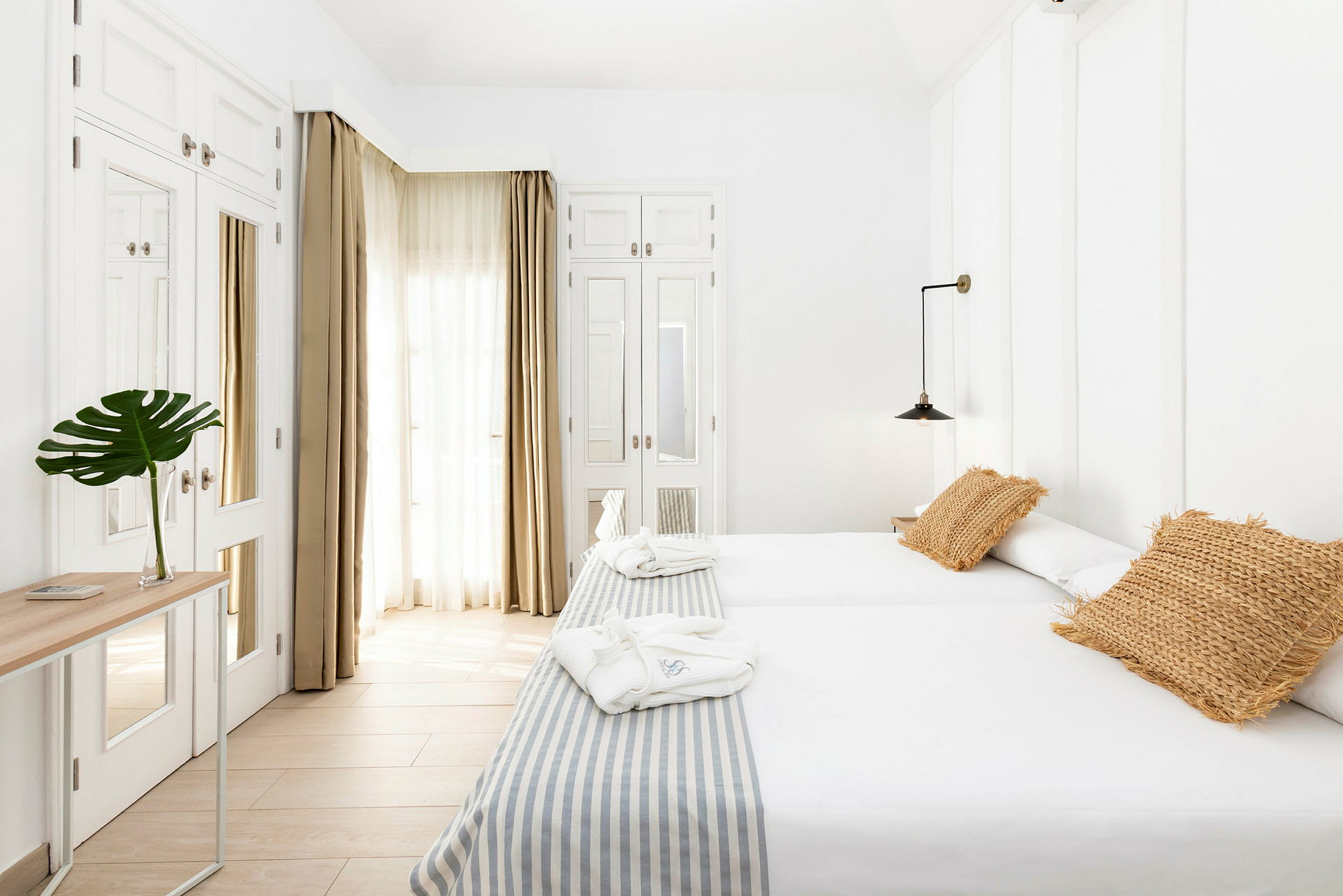 Valoisa hotellihuone, jossa kaksi erillistä sänkyä, beigejä tyynyjä ja pöytä, jossa vihreä kasvi lasimaljakossa. (Tekoälyn luoma teksti)