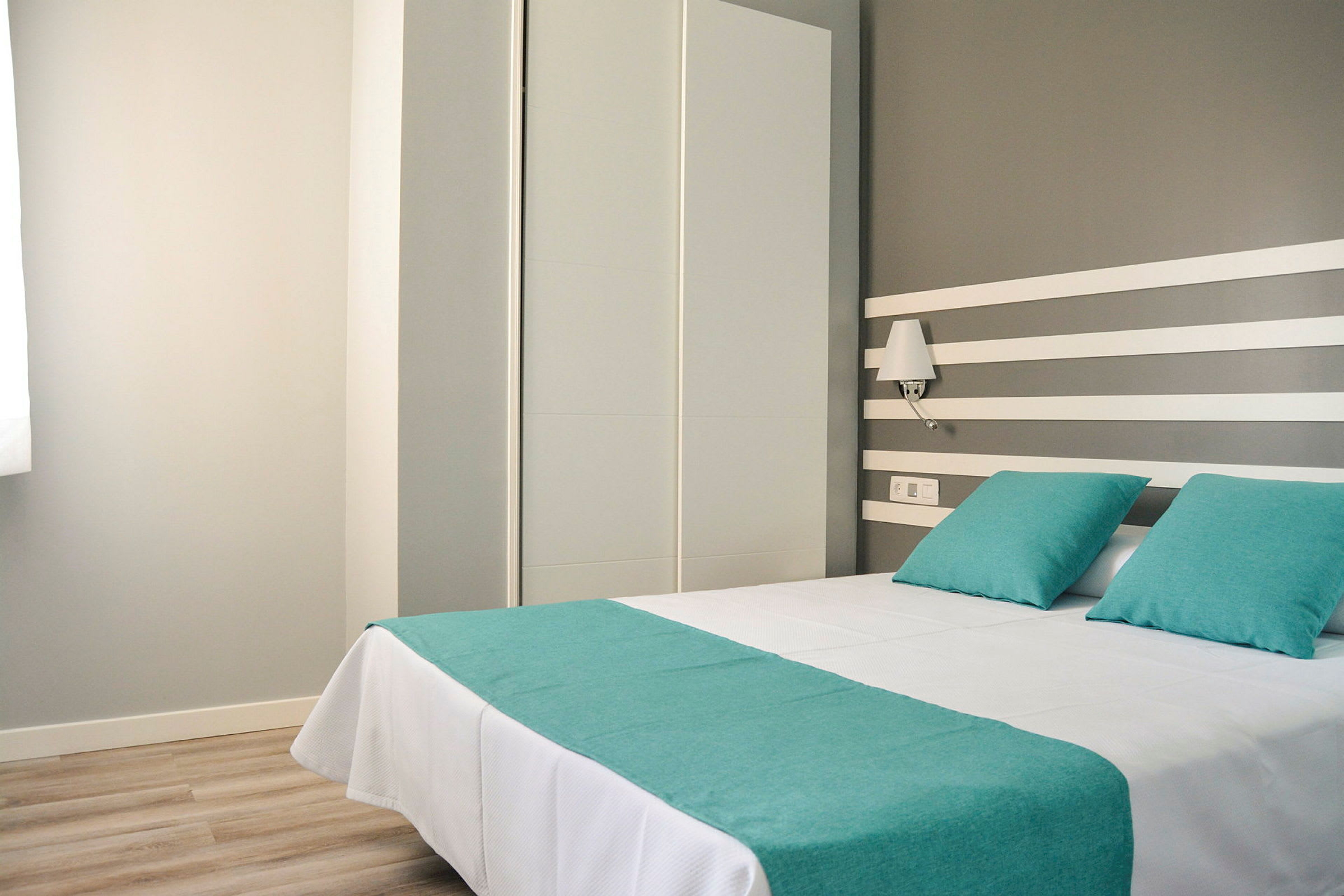 Hotellihuone, jossa on parivuode, peitetty valkoisilla lakanoilla ja turkooseilla tyynyillä sekä päiväpeitteellä. (Tekoälyn luoma teksti)