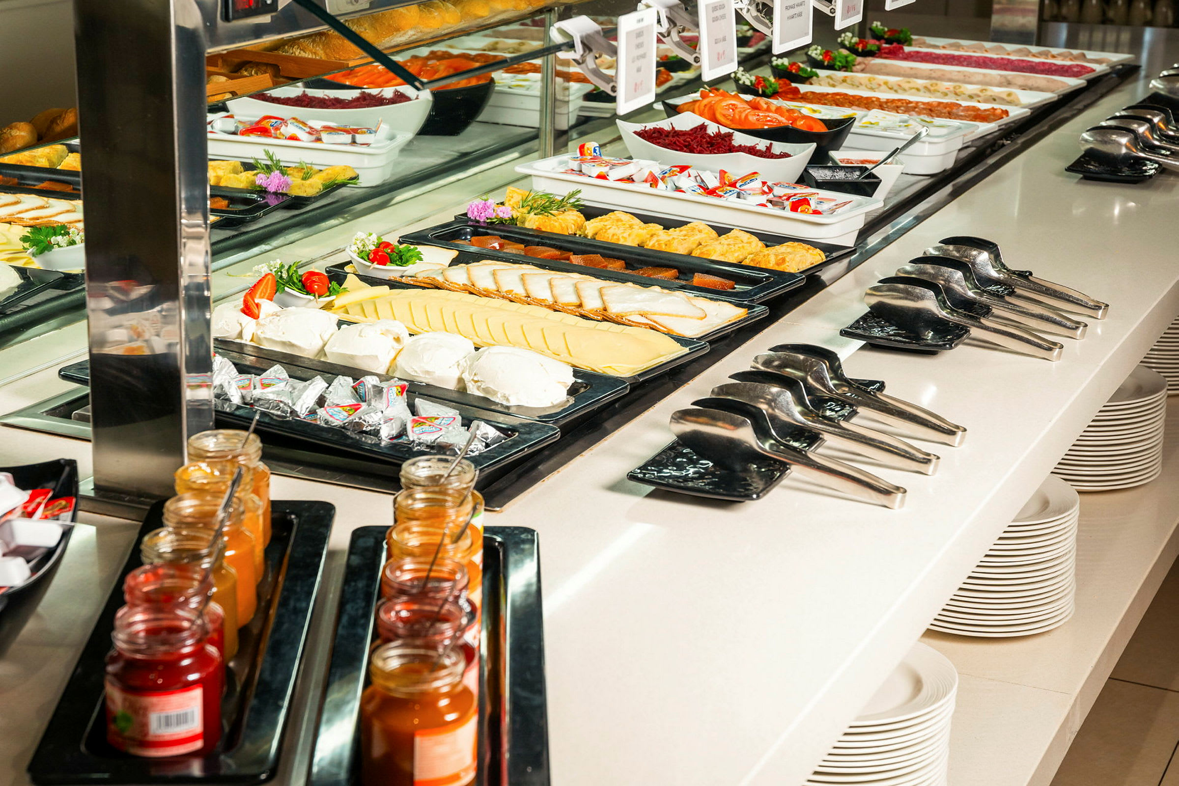 Buffet, jossa on erilaisia ruokia, kuten juustosiivuja, vihanneksia ja hilloja purkeissa, tarjoiluvälineitä ja pinottuja lautasia. (Tekoälyn luoma teksti)