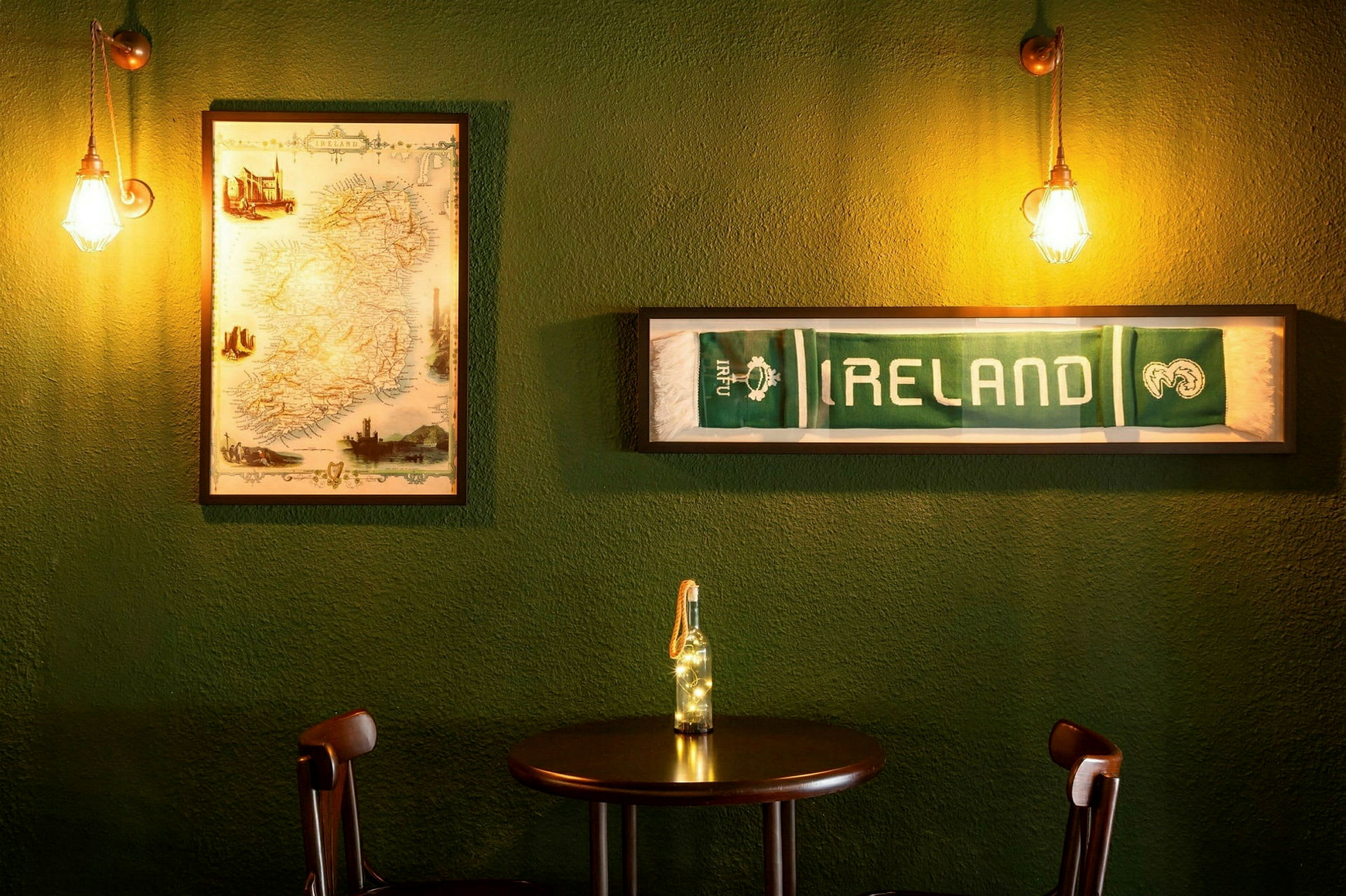 Pöytä ja kaksi tuolia vihreän seinän edessä, jota koristavat kehystetty Irlannin kartta ja kehystetty huivi tekstillä "IRELAND". (Tekoälyn luoma teksti)