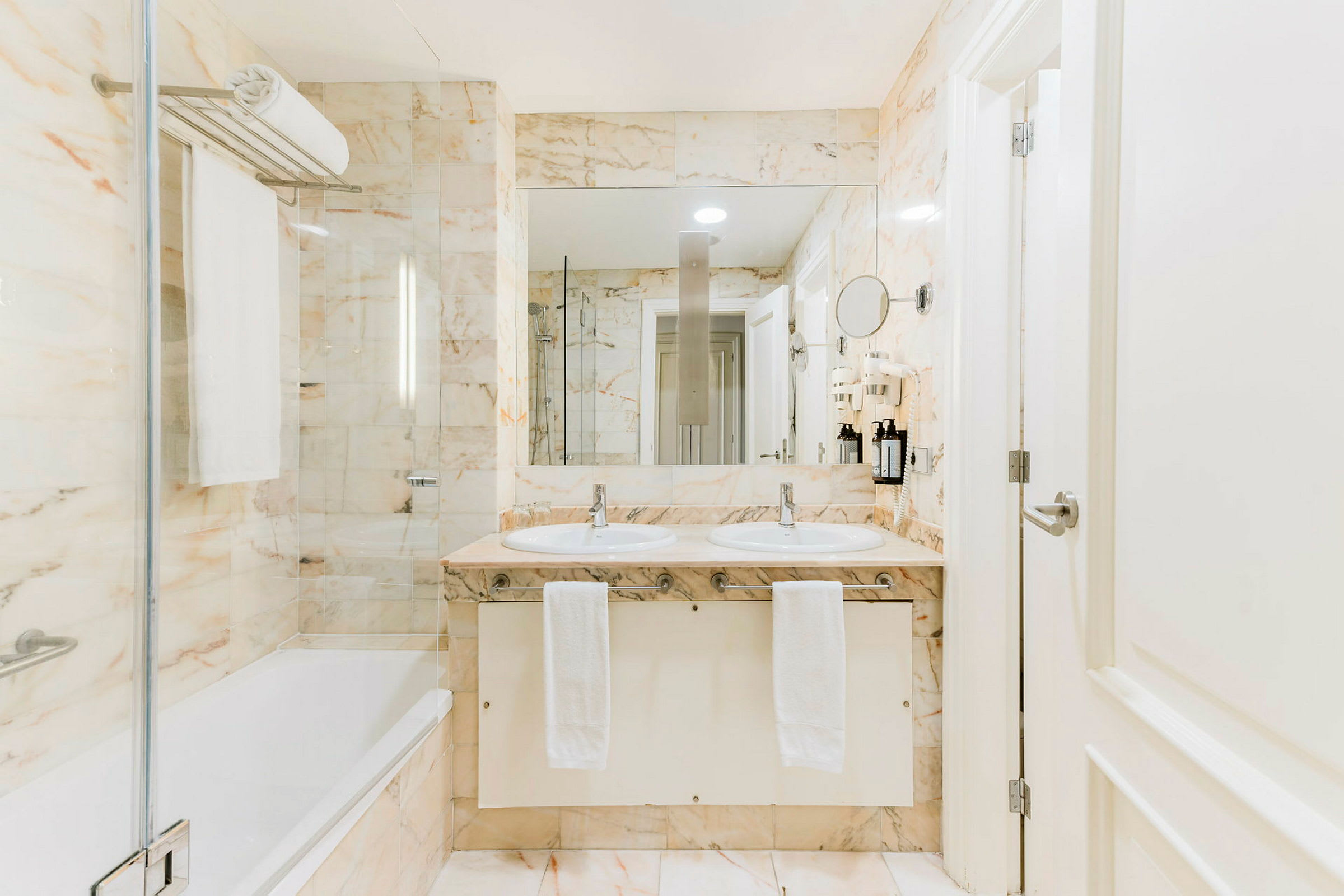 Vaalea kylpyhuone, jossa on kaksi pesuallasta, suuri peili ja kylpyamme, marmorikuvioiset seinät ja lattia. (Tekoälyn luoma teksti)