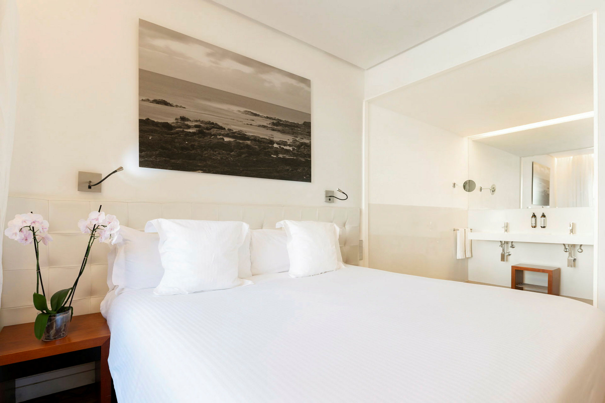Hotellihuone, jossa on pedattu sänky etualalla, valkoiset vuodevaatteet ja kaksi orkideaa yöpöydällä vieressä. Taustalla näkyy kylpyhuone kahdella pesualtaalla. (Tekoälyn luoma teksti)