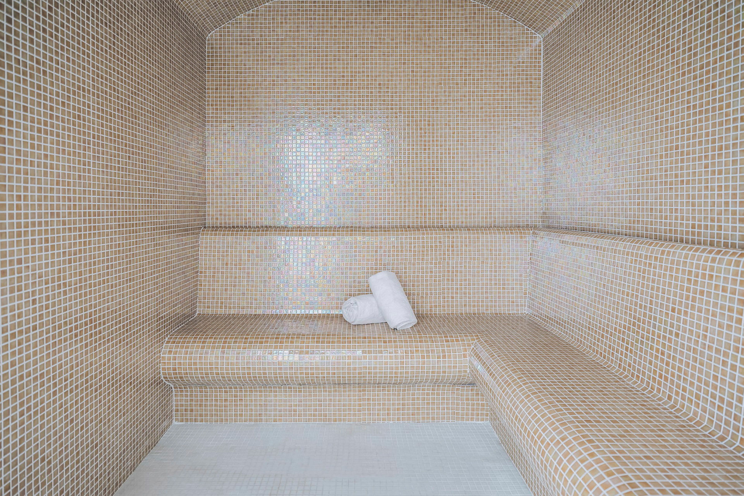 Höyryhuone, jossa seinät ja penkit on päällystetty pienillä beige-mosaiikkilaatoilla. Kaksi valkoista pyyhettä on asetettu penkille. (Tekoälyn luoma teksti)