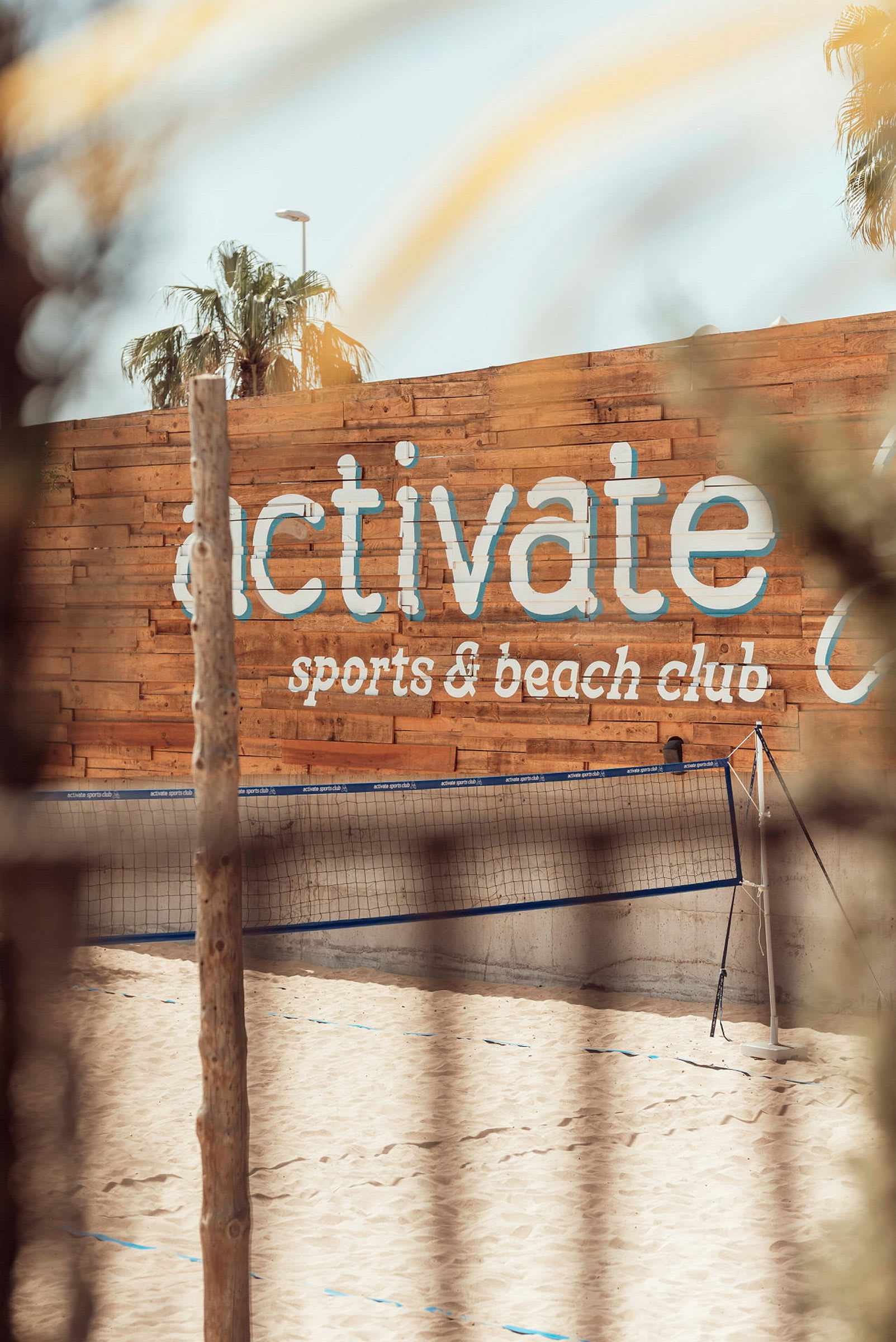 Hiekkaranta ja lentopalloverkko, taustalla puuseinä, jossa lukee 'activate sports & beach club'. (Tekoälyn luoma teksti)