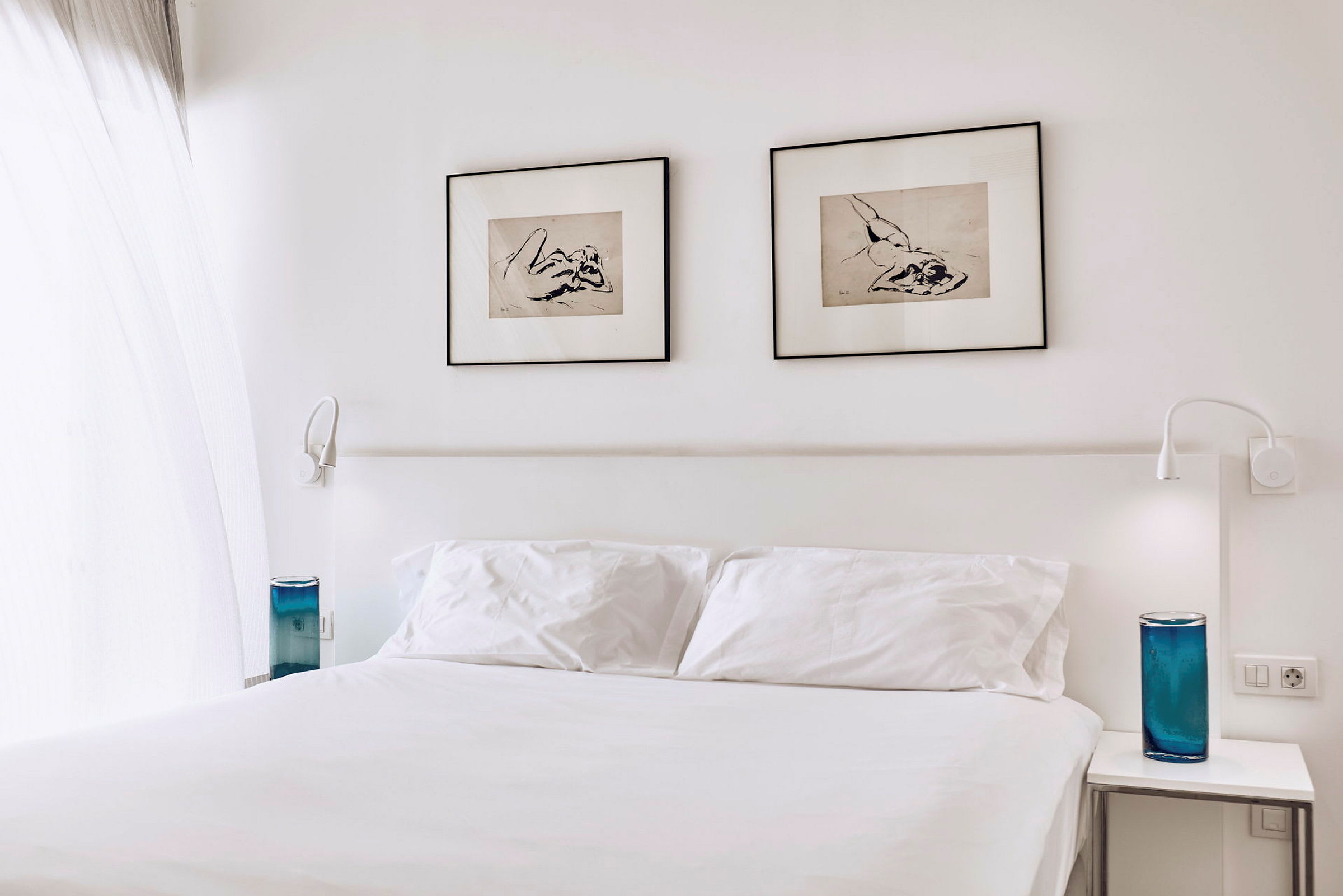 Hotellihuone, jossa on valkoinen parisänky, kaksi kehystettyä taideteosta seinällä ja siniset koriste-esineet yöpöydillä. (Tekoälyn luoma teksti)