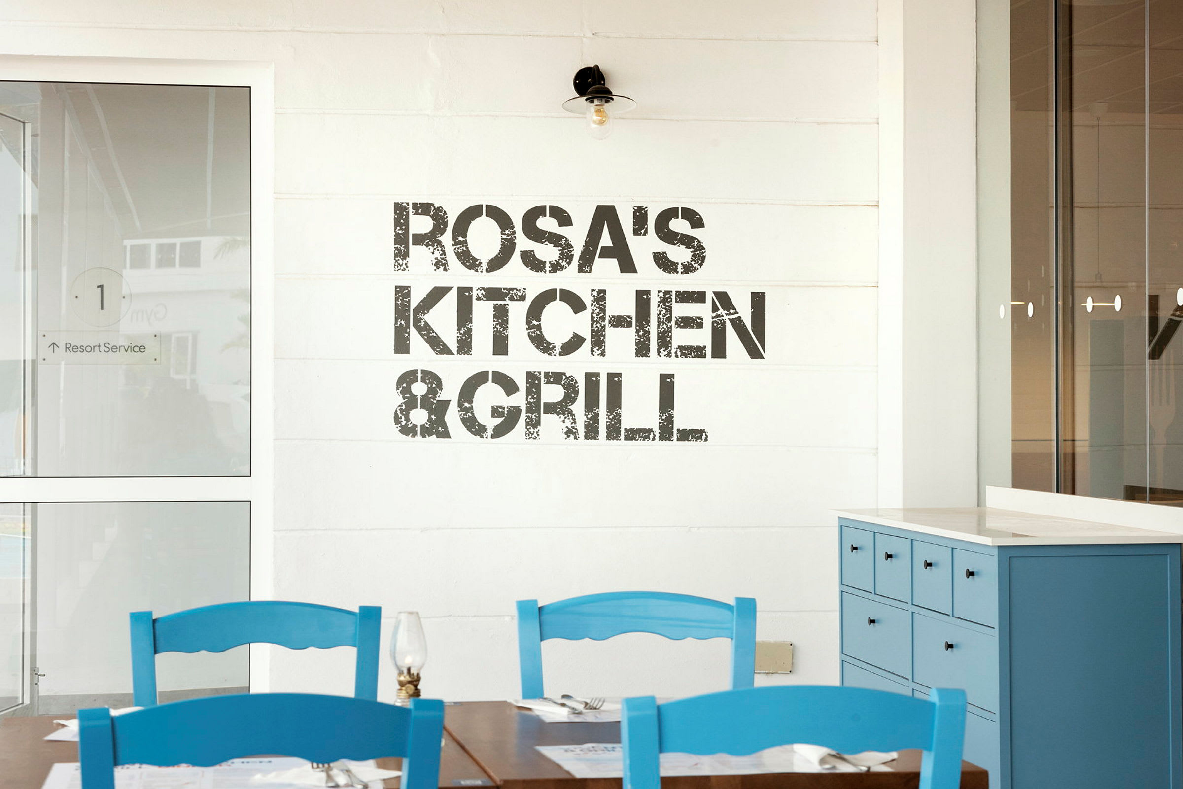 Kyltti, jossa lukee 'Rosa's Kitchen & Grill', seinällä, etualalla pöytiä ja sinisiä tuoleja. (Tekoälyn luoma teksti)