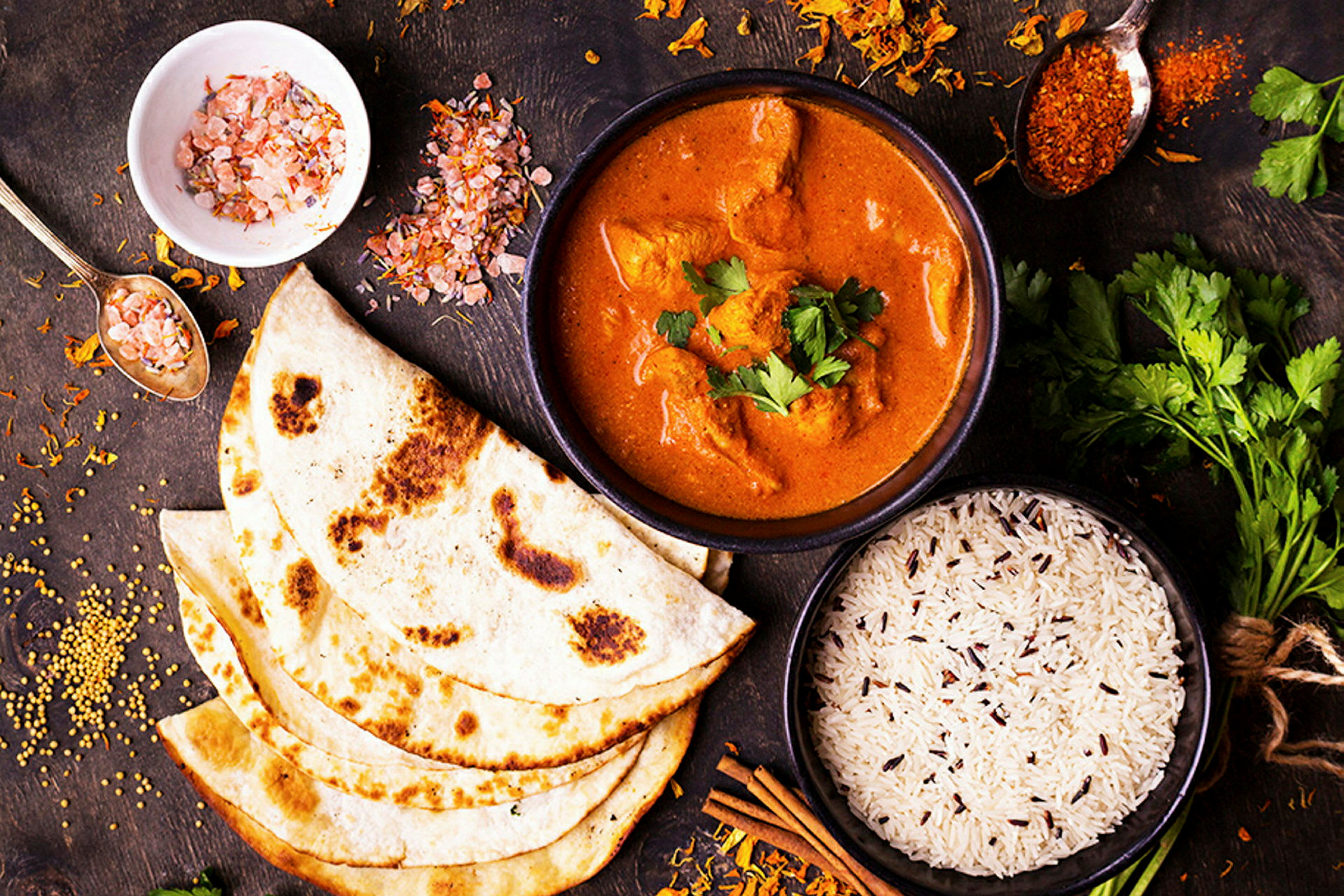 Asetelma, jossa on mausteinen currypata, riisiä ja naan-leipää sekä mausteita ja yrttejä puupöydällä. (Tekoälyn luoma teksti)