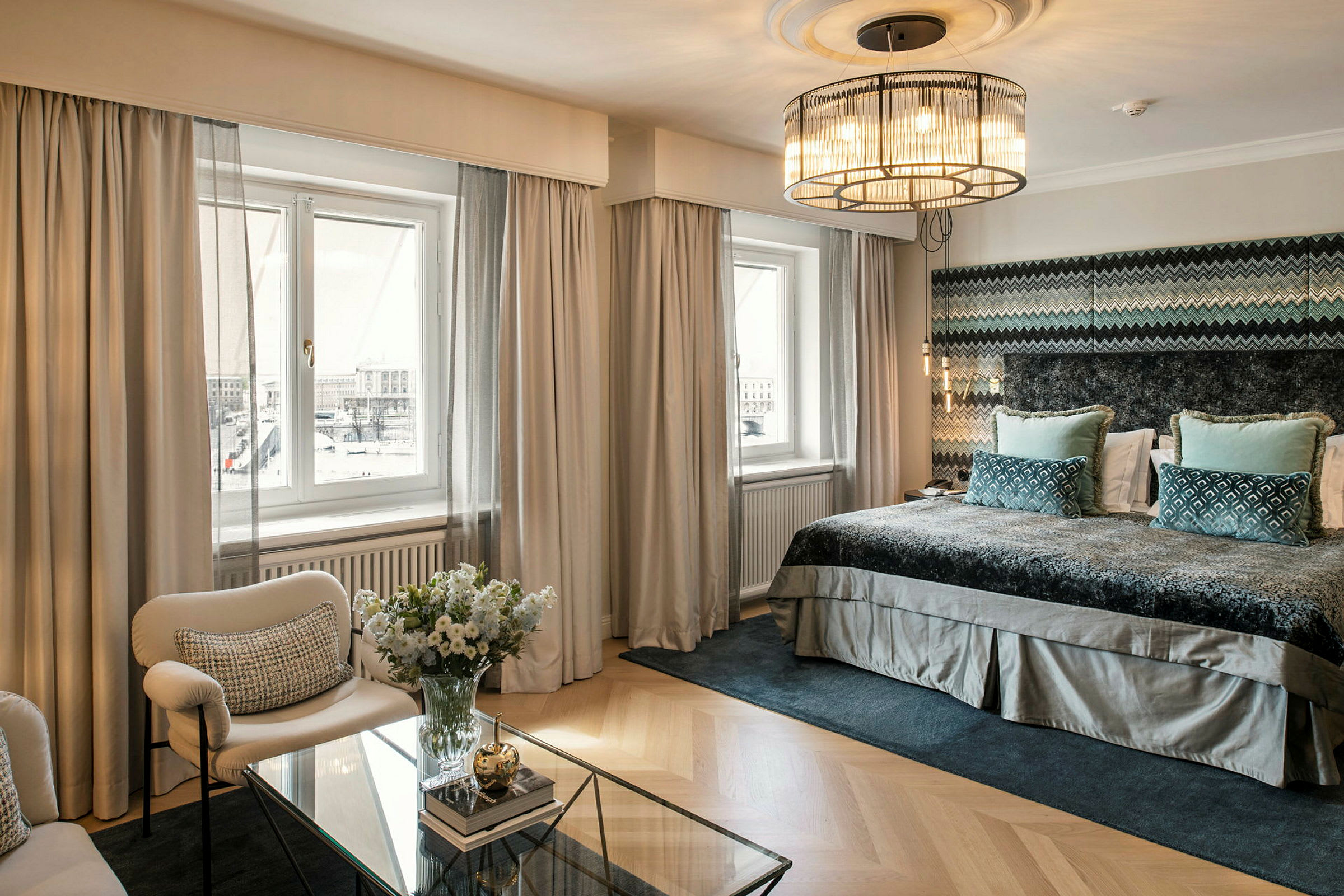 Tyylikäs hotellihuone, jossa on suuret ikkunat, leveä sänky, mukavat istuimet ja lasipöytä koristeellisella somistuksella. (Tekoälyn luoma teksti)