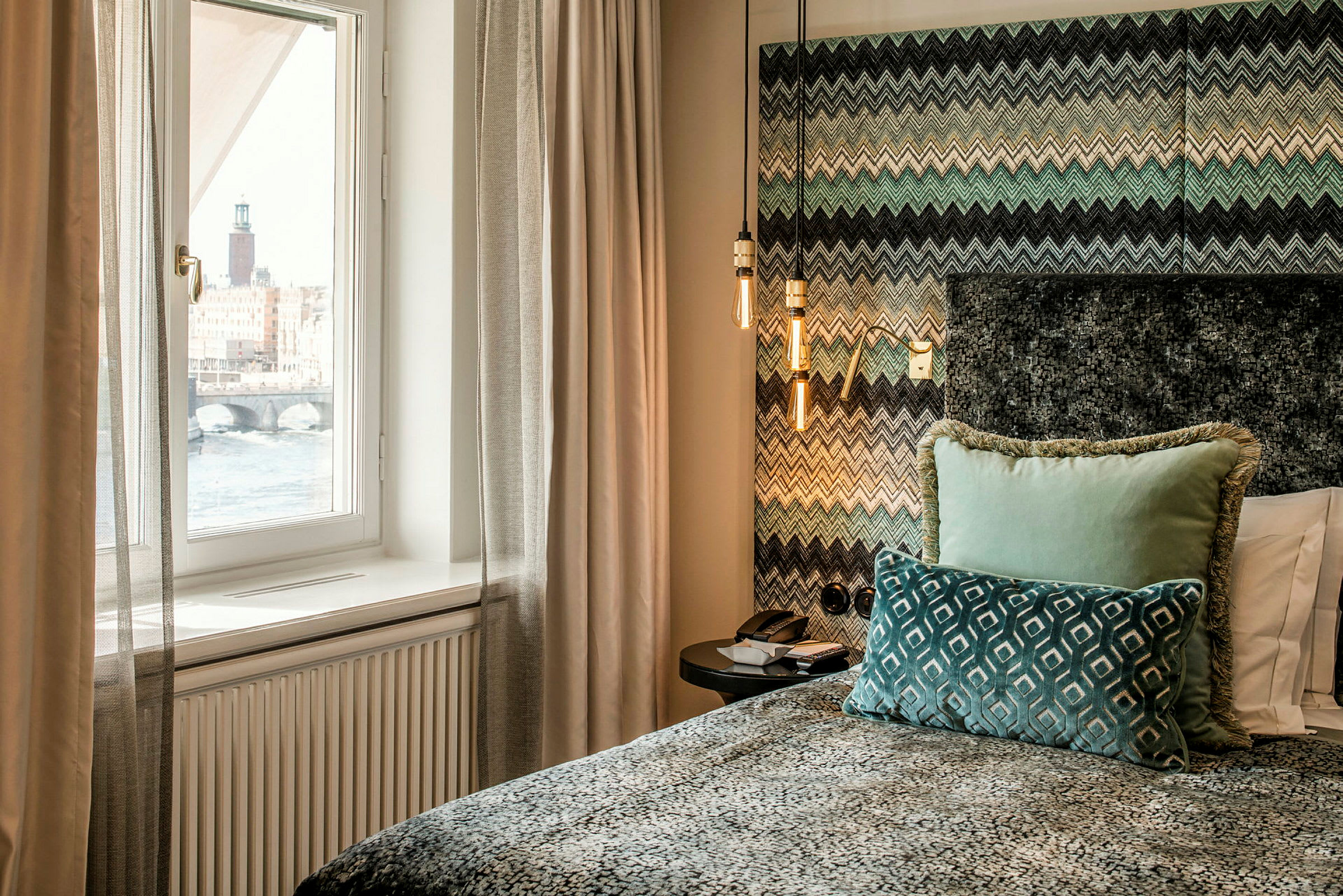 Moderni hotellihuone, jossa ikkunasta näkyy vesistö ja kaupungintalo. Sänky on koristeltu värikkäillä tyynyillä ja kuvioidulla peitteellä. (Tekoälyn luoma teksti)