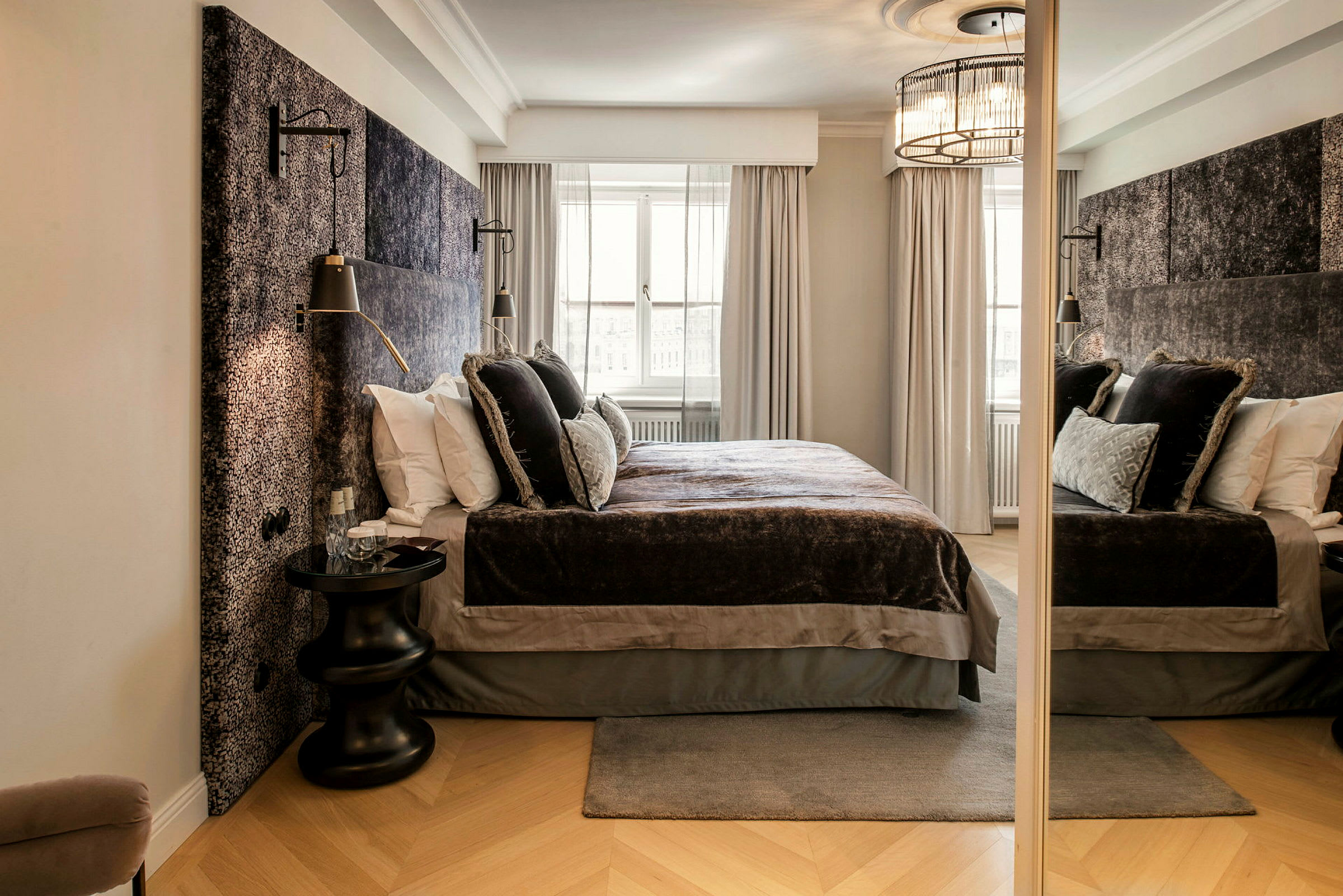 Tyylikäs hotellihuone, jossa on suuri sänky pehmeillä päiväpeitteillä ja koristeellisilla tyynyillä. Huoneessa on vaaleat verhot ikkunan edessä ja sivulla peili. (Tekoälyn luoma teksti)