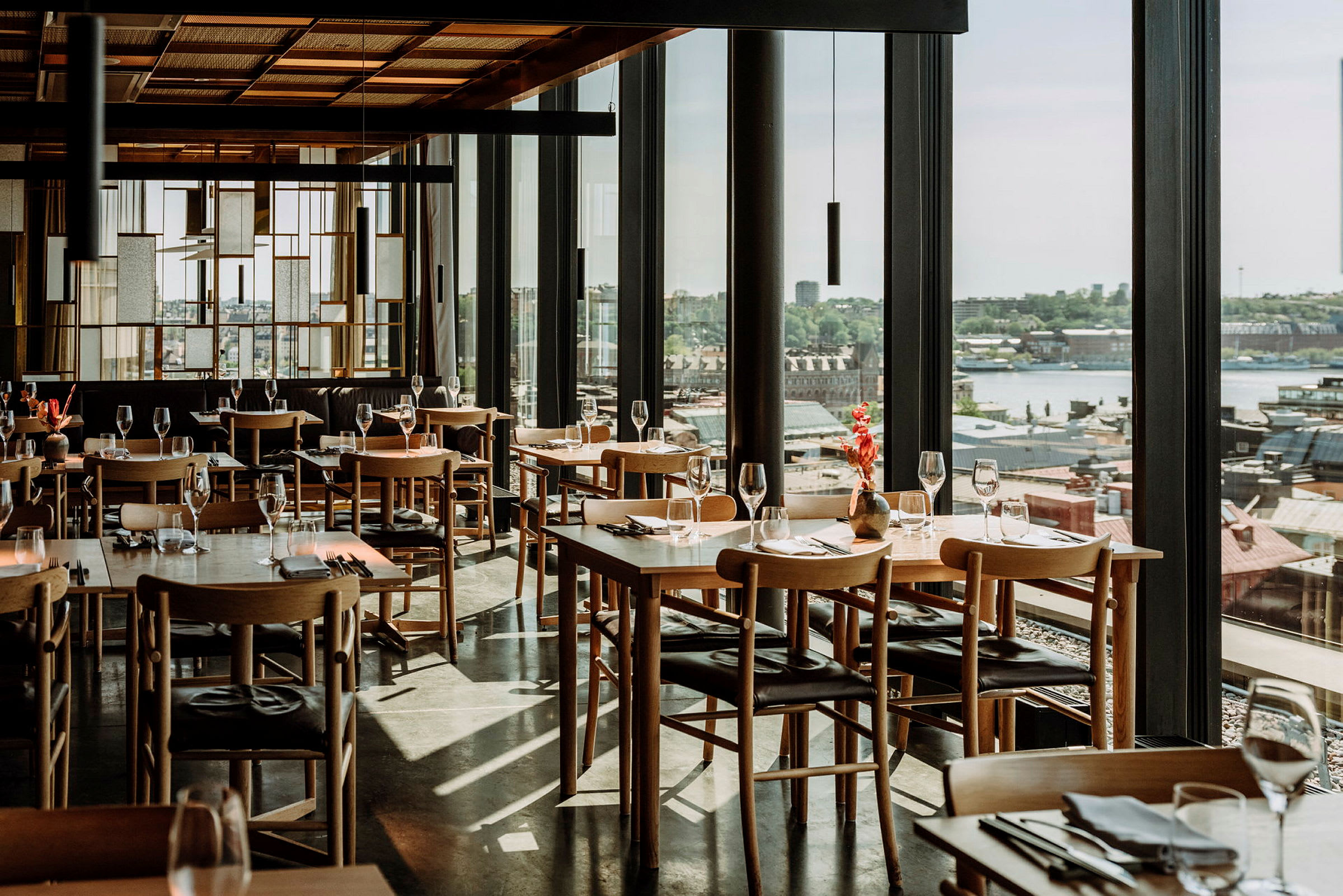 Tyylikäs ravintola, jossa on puupöydät ja -tuolit, lasia heijastamassa valoa sekä panoraamanäkymä kaupunkiin. (Tekoälyn luoma teksti)
