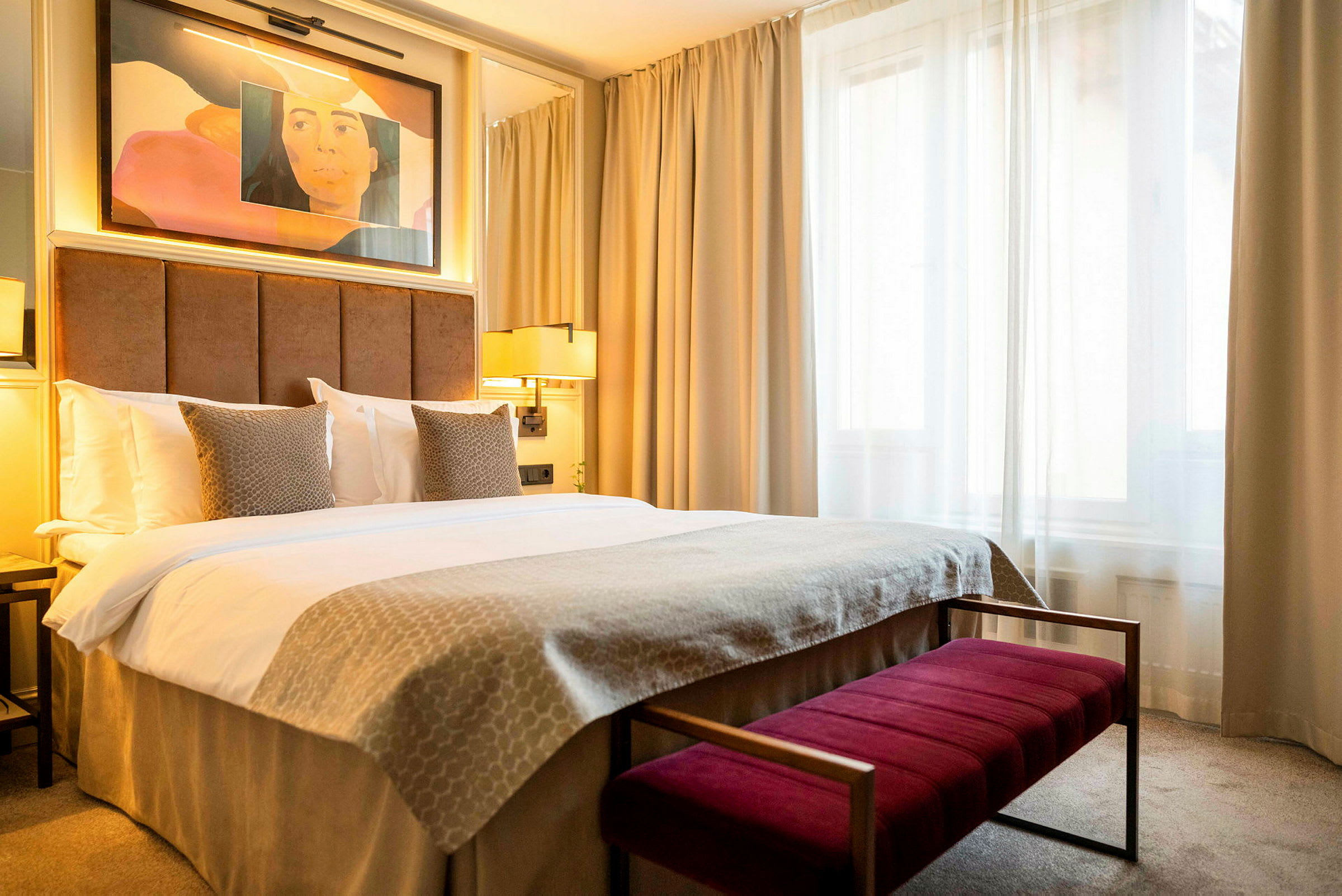 Hotellihuone, jossa on parivuode, beige vuodevaatteet ja kaksi tyynyä pehmustetun ruskean sängynpäädyn edessä. Taustalla näkyy valoja ja ikkuna vaaleilla verhoilla. (Tekoälyn luoma teksti)