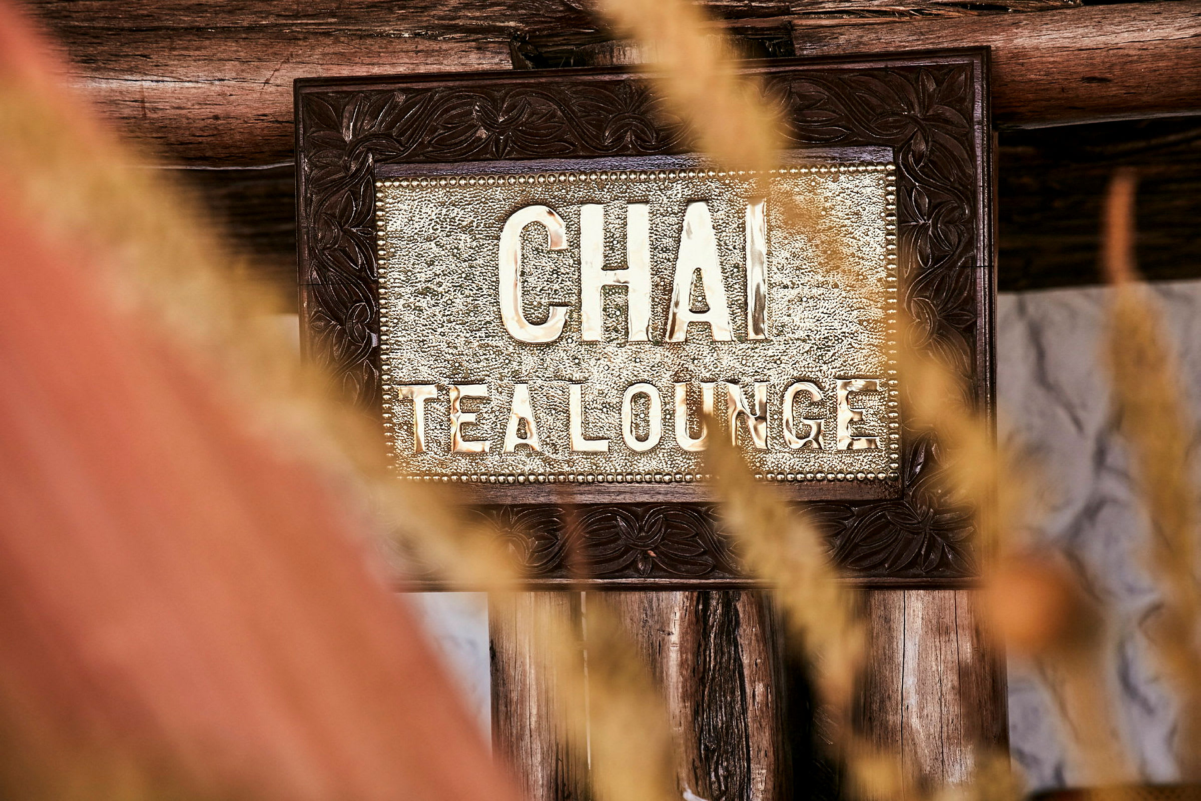Lähikuva kyltistä, jossa lukee 'Chai Tea Lounge', ympäröitynä puukoristeilla ja kuihtuneella ruoholla etualalla. (Tekoälyn luoma teksti)