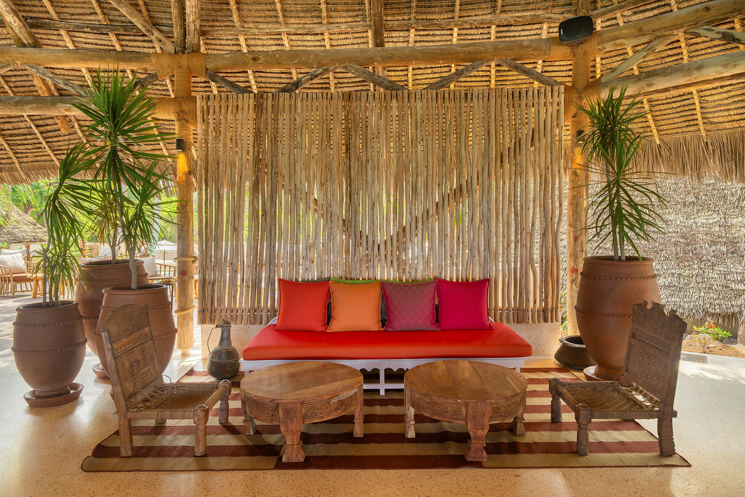 Taukotila, jossa on oksista ja oljista tehty katto, punainen sohva koristeellisilla värikkäillä tyynyillä, rustiikkisia puukalusteita ja suuria keraamisia ruukkuja vihreillä kasveilla. (Tekoälyn luoma teksti)