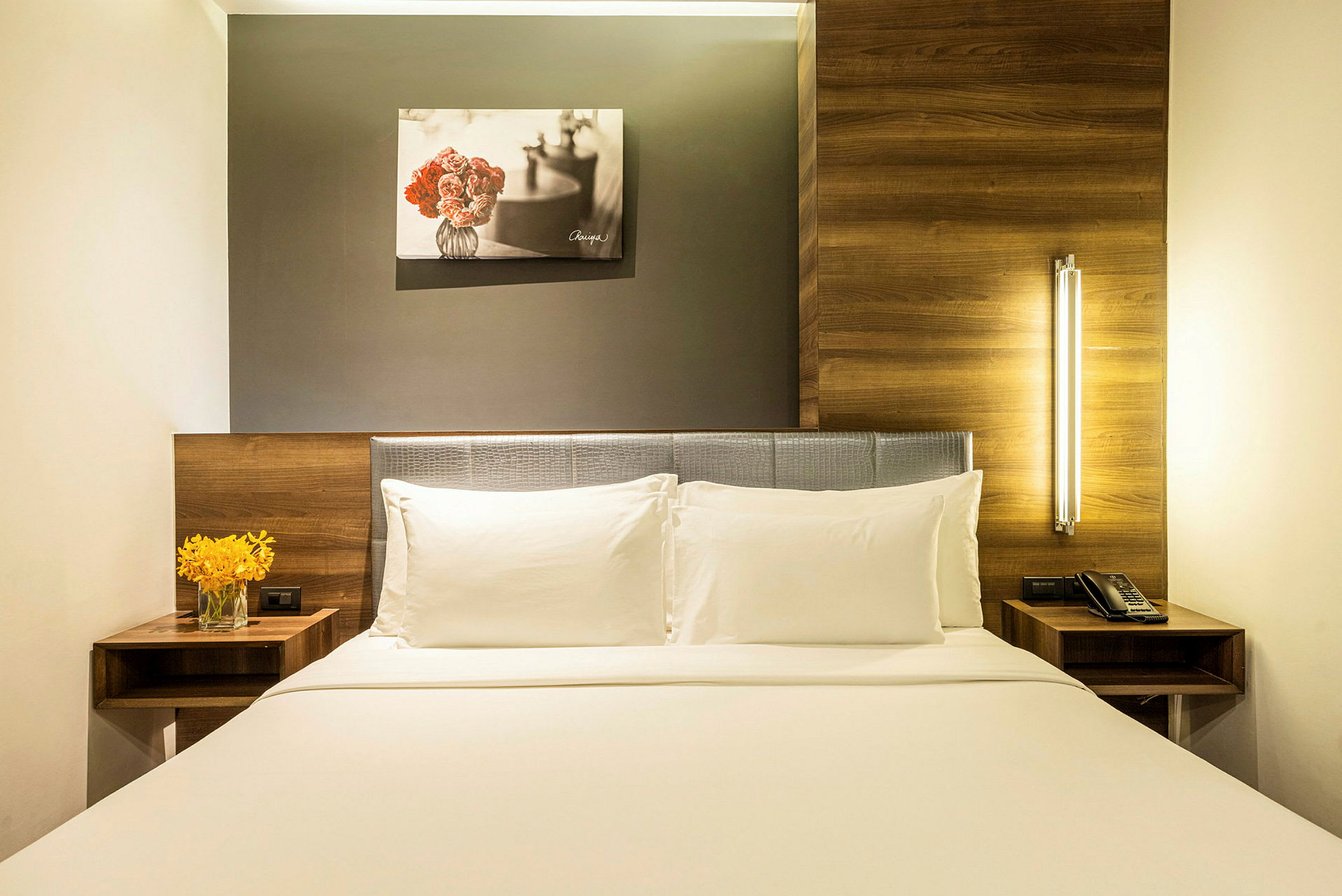 Hotellihuone, jossa on mukava parisänky, kaksi valkoista tyynyä ja valkoinen vuodevaatekangas. Yöpöydällä vasemmalla on maljakko, jossa on keltaisia kukkia, ja oikealla toinen yöpöytä, jossa on puhelin. Sängyn yläpuolella on kukkamaalaus. (Tekoälyn luoma teksti)
