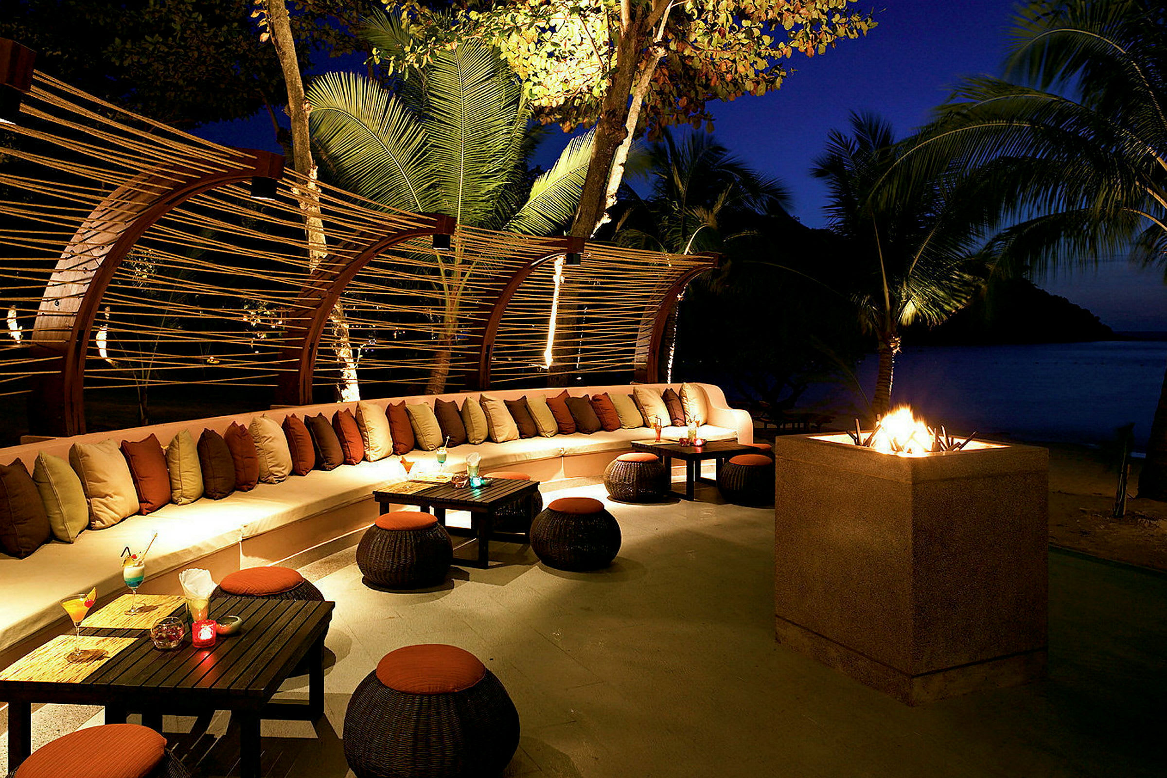 Ulkoalue, jossa on pitkä valkoinen sohva täynnä tyynyjä, pieniä pöytiä ympärillä kynttiläkoristeilla sekä avoin tulisija. Trooppinen ympäristö, jota ympäröivät palmut. (Tekoälyn luoma teksti)