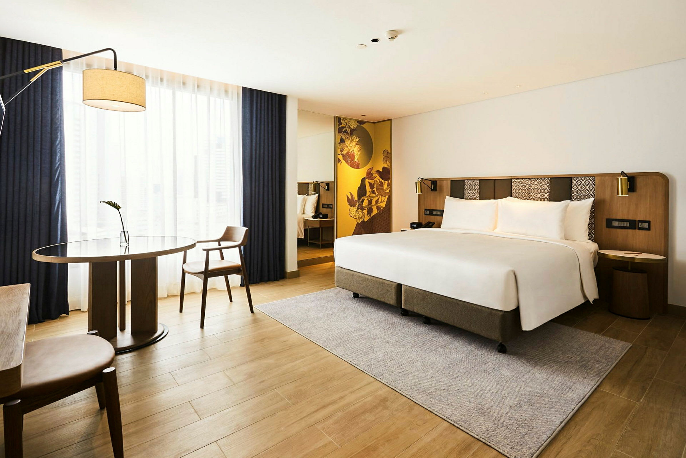 Moderni hotellihuone, jossa on parivuode, pöytä ja kaksi tuolia sekä suuri ikkunapinta verhoilla. (Tekoälyn luoma teksti)