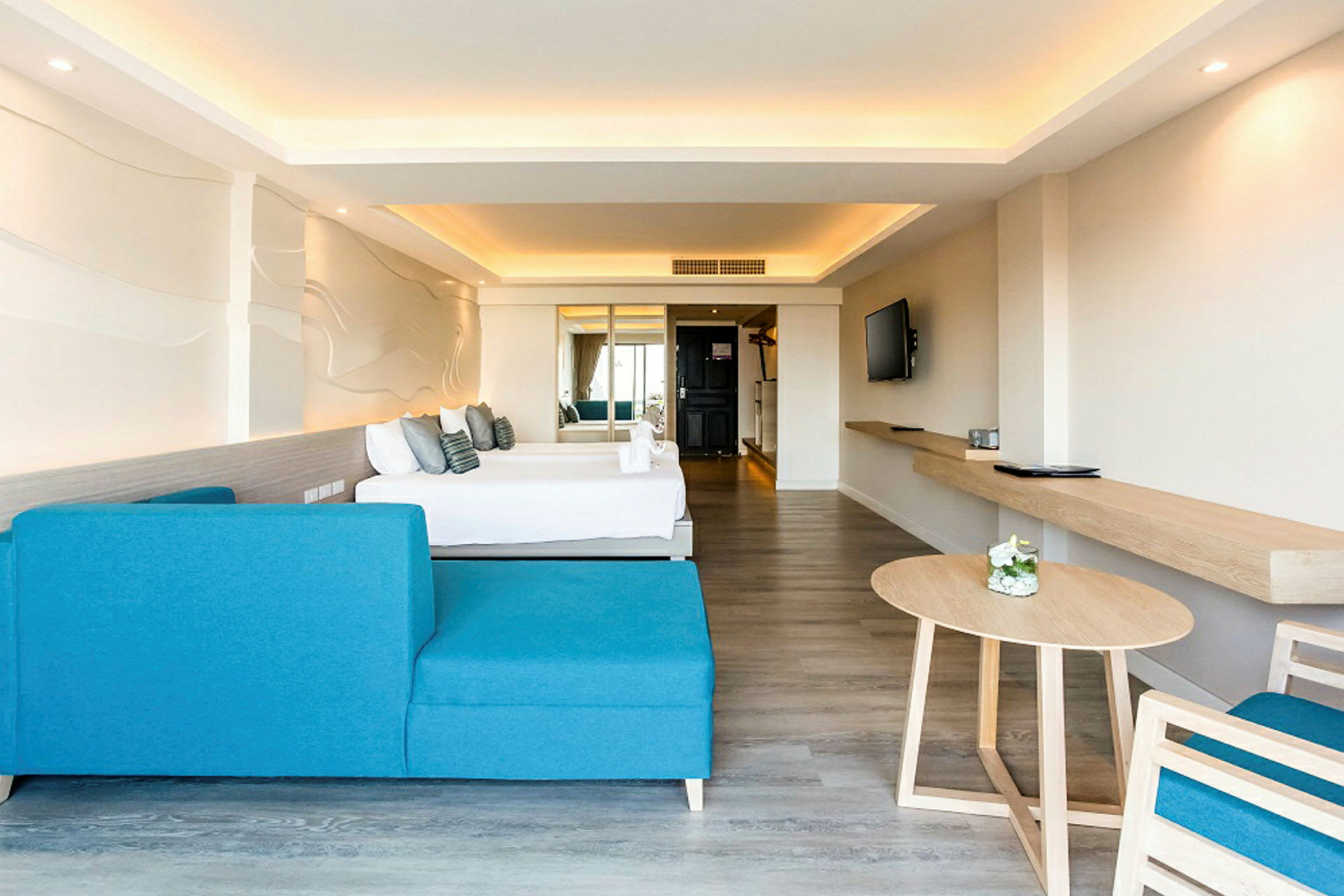 Moderni hotellihuone, jossa on sininen sohva, pöytä ja taustalla sänky. Seinällä on koristeltu kuvio, ja huone on valoisa neutraalein värein. (Tekoälyn luoma teksti)