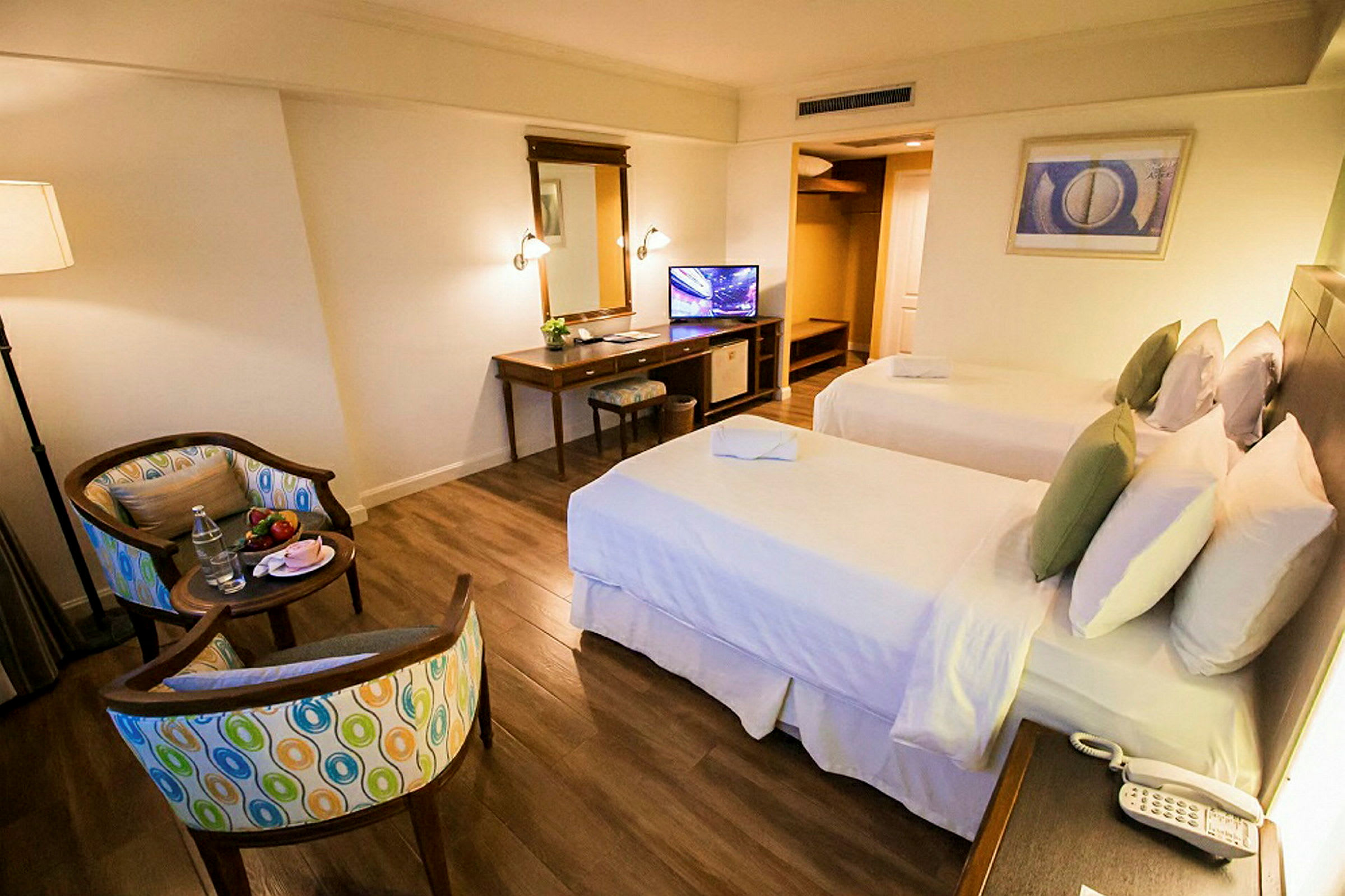 Hotellihuone, jossa on kaksi erillistä sänkyä, kirjoituspöytä, tuoli, peili ja televisio. Huoneessa on puulattia ja istuinryhmä, johon kuuluu kaksi tuolia ja pieni pöytä. (Tekoälyn luoma teksti)