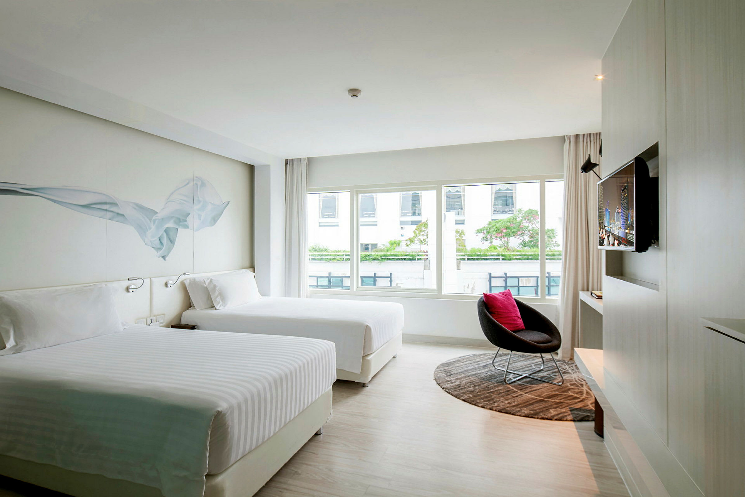 Moderni hotellihuone, jossa on kaksi suurta sänkyä, iso ikkunaseinä näkymällä, seinälle asennettu televisio ja tuoli pyöreän maton päällä. (Tekoälyn luoma teksti)