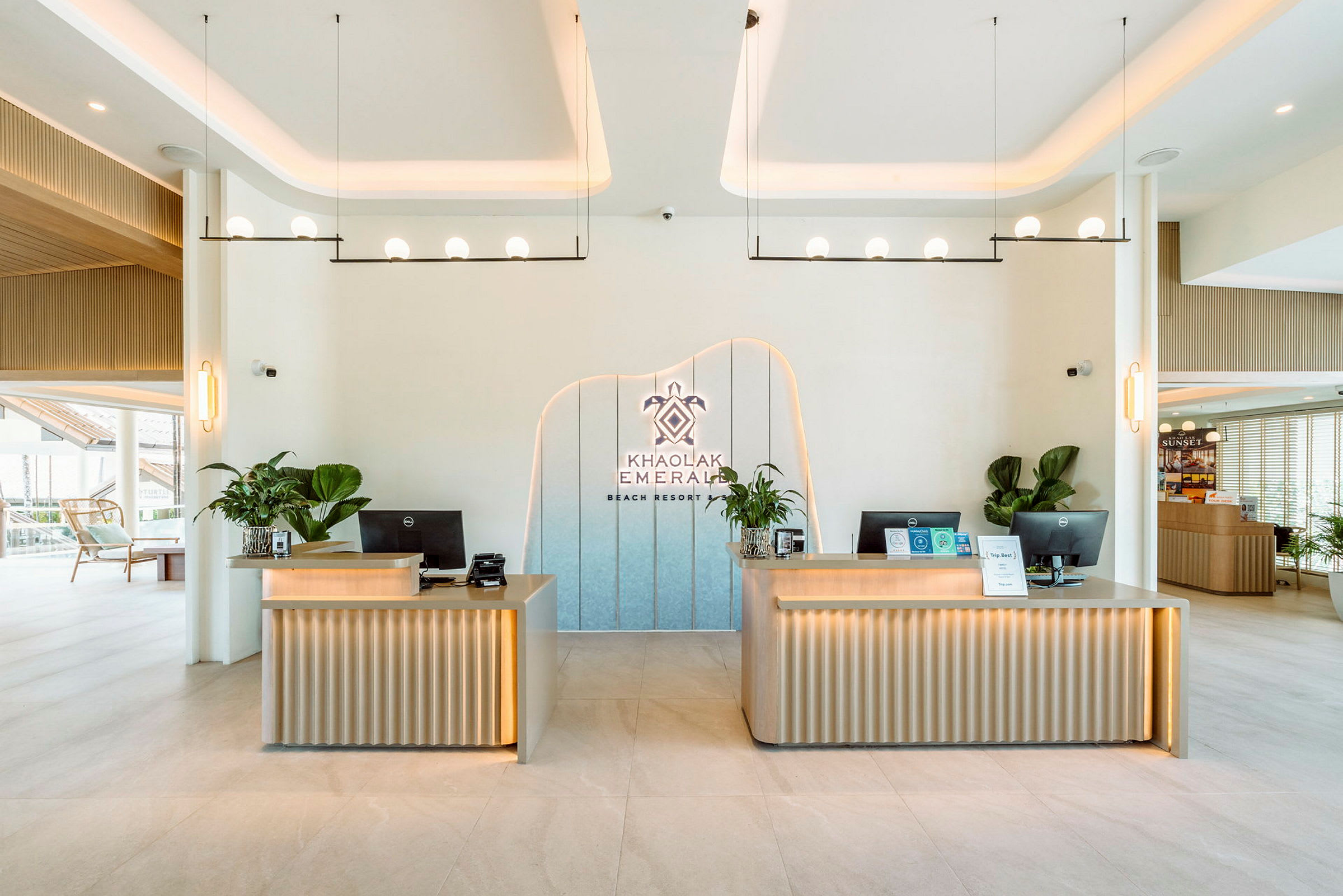 Vastaanottoalue, jossa kaksi modernia pöytää koristeena kasveja. Seinälle on sijoitettu logo, jossa lukee 'Khao Lak Emerald Beach Resort'. (Tekoälyn luoma teksti)