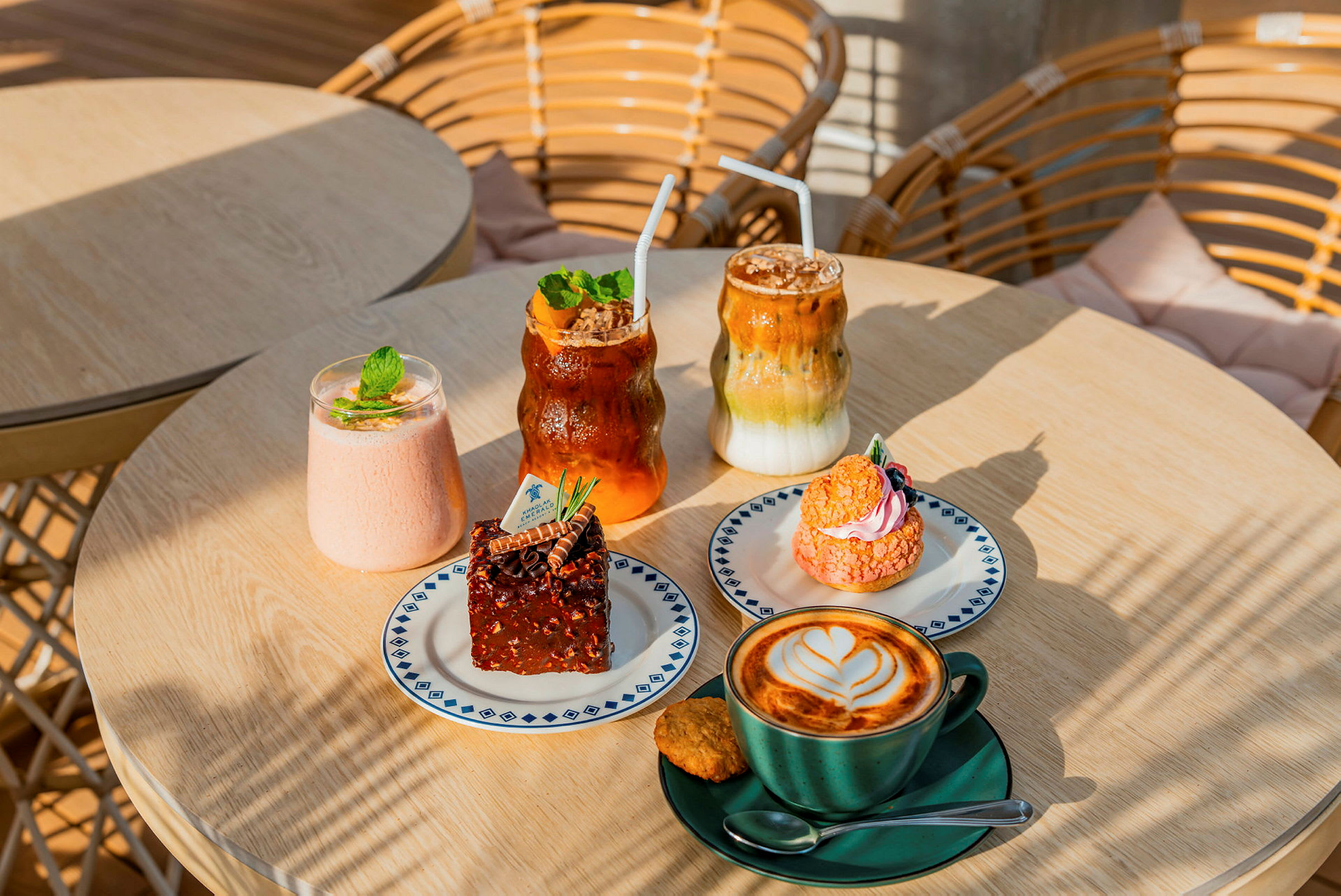 Kuvassa on pöytä, jossa on erilaisia leivonnaisia ja juomia. Siinä on latteartilla koristeltu kahvikuppi, brownie, vaaleanpunainen leivonnainen ja muutama kylmä juoma. (Tekoälyn luoma teksti)