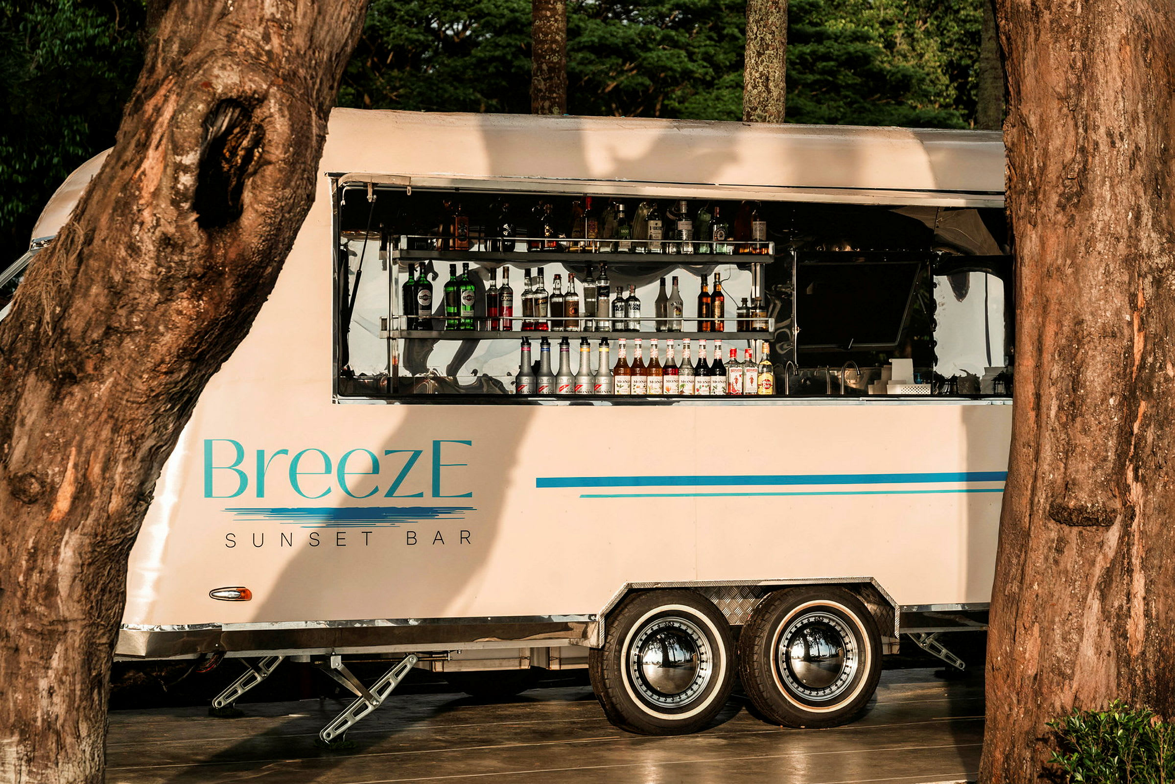 Liikkuva baaripiste, jossa on merkintä 'BreezE Sunset Bar', on asetettu puiden väliin, ja siinä on näkyvillä pulloja ja juomia avoimessa palvelualueessa. (Tekoälyn luoma teksti)