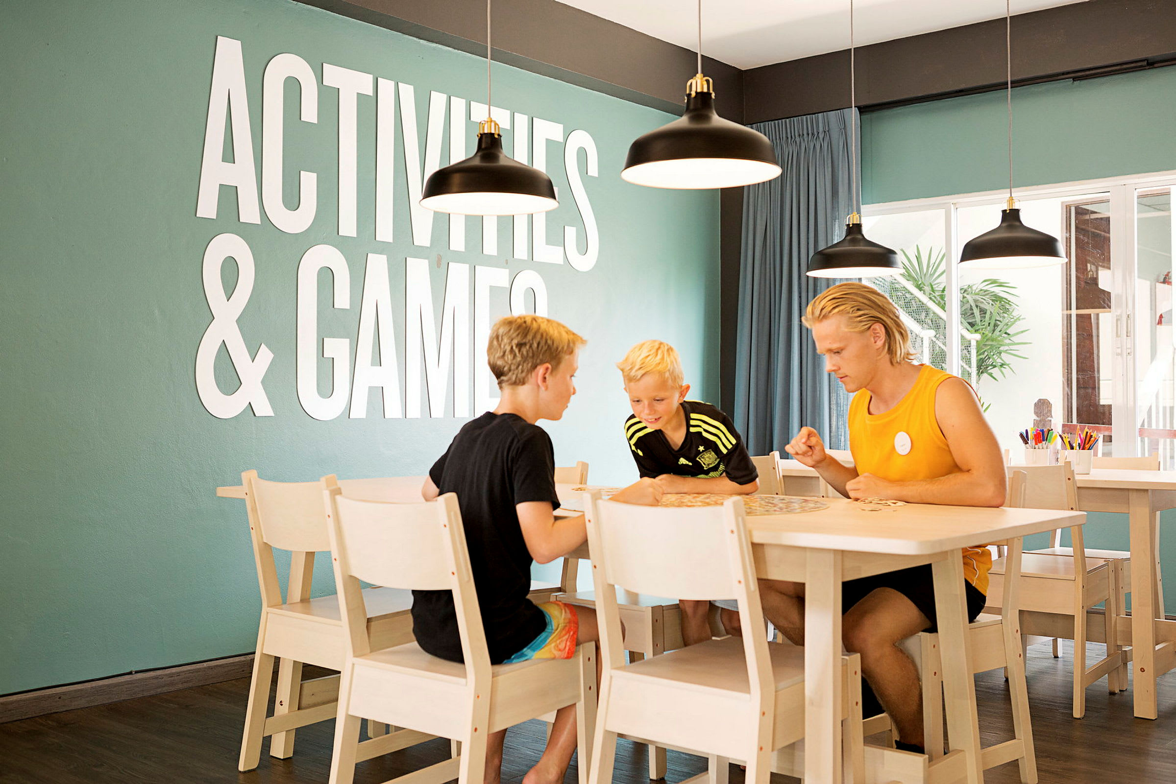 Henkilöt pelaavat lautapeliä pöydän ääressä huoneessa, jonka seinällä lukee "Activities & Games". (Tekoälyn luoma teksti)