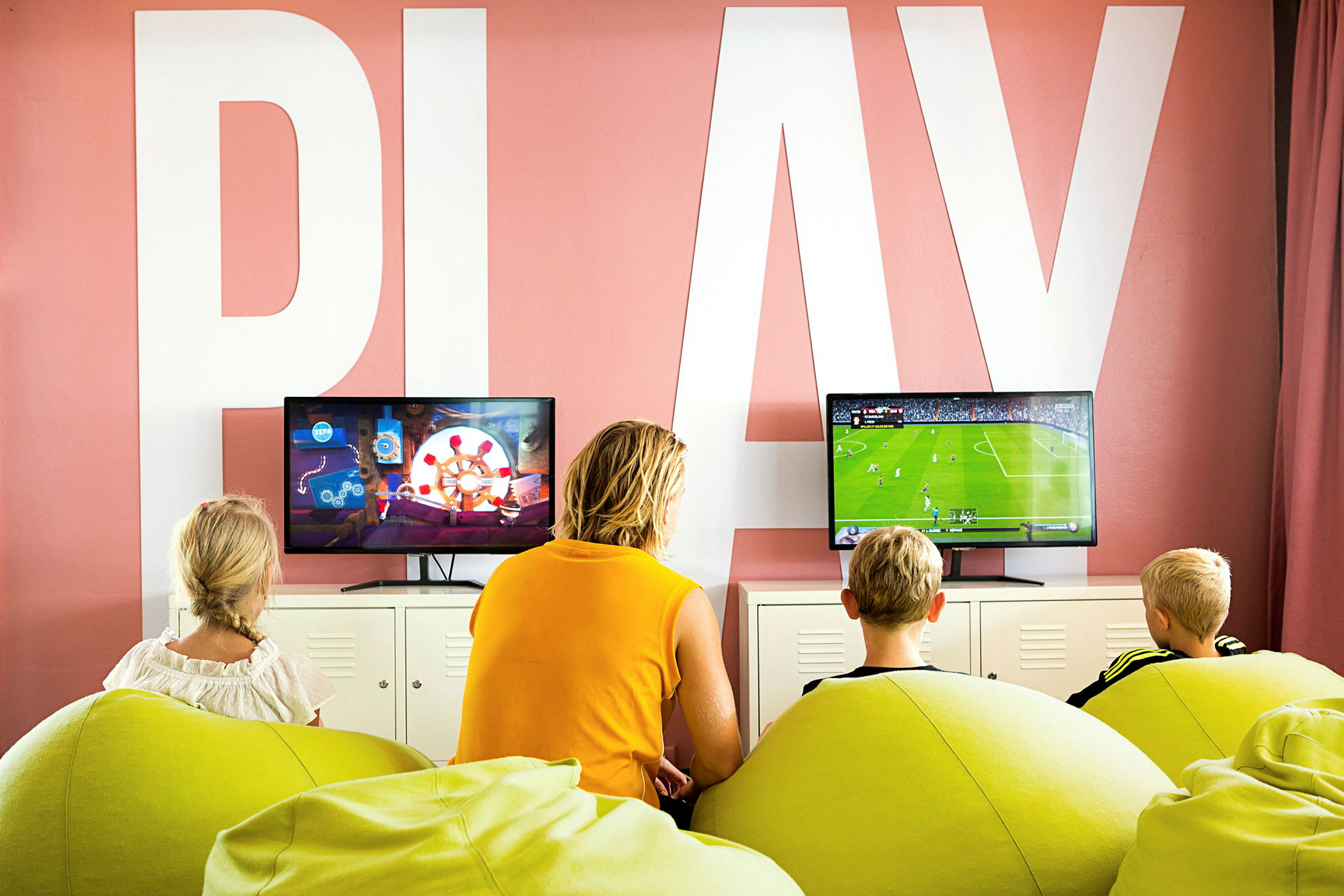 Kolme lasta ja yksi aikuinen istuvat vihreillä säkkituoleilla pelaamassa televisiopelejä kahden näytön edessä huoneessa, jossa seinällä lukee 'PLAY'. (Tekoälyn luoma teksti)