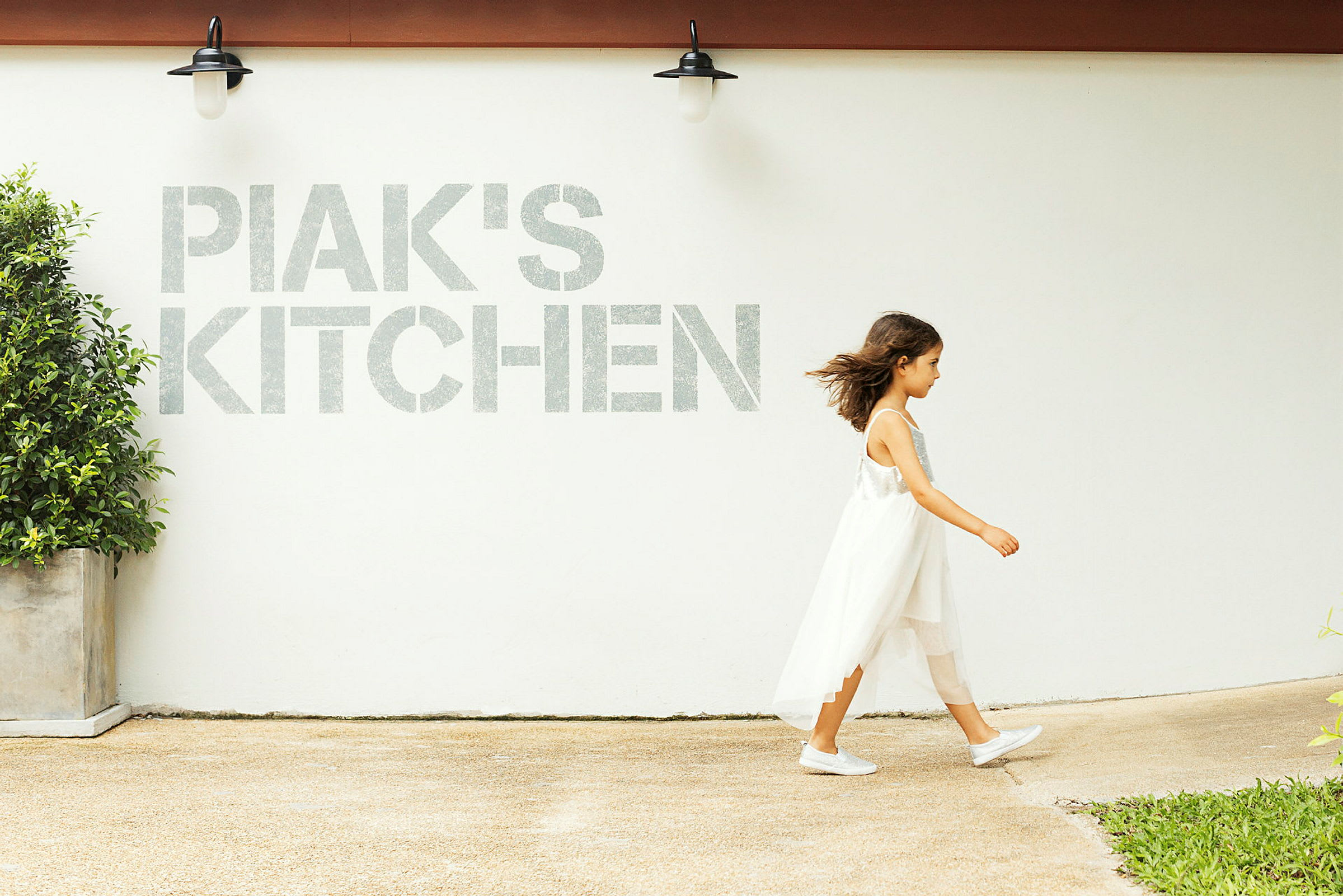 Henkilö kävelee valkoisen seinän ohi, jossa lukee 'PIAK'S KITCHEN', ja näkyvissä on lamppu sekä ruukkukasvi. (Tekoälyn luoma teksti)
