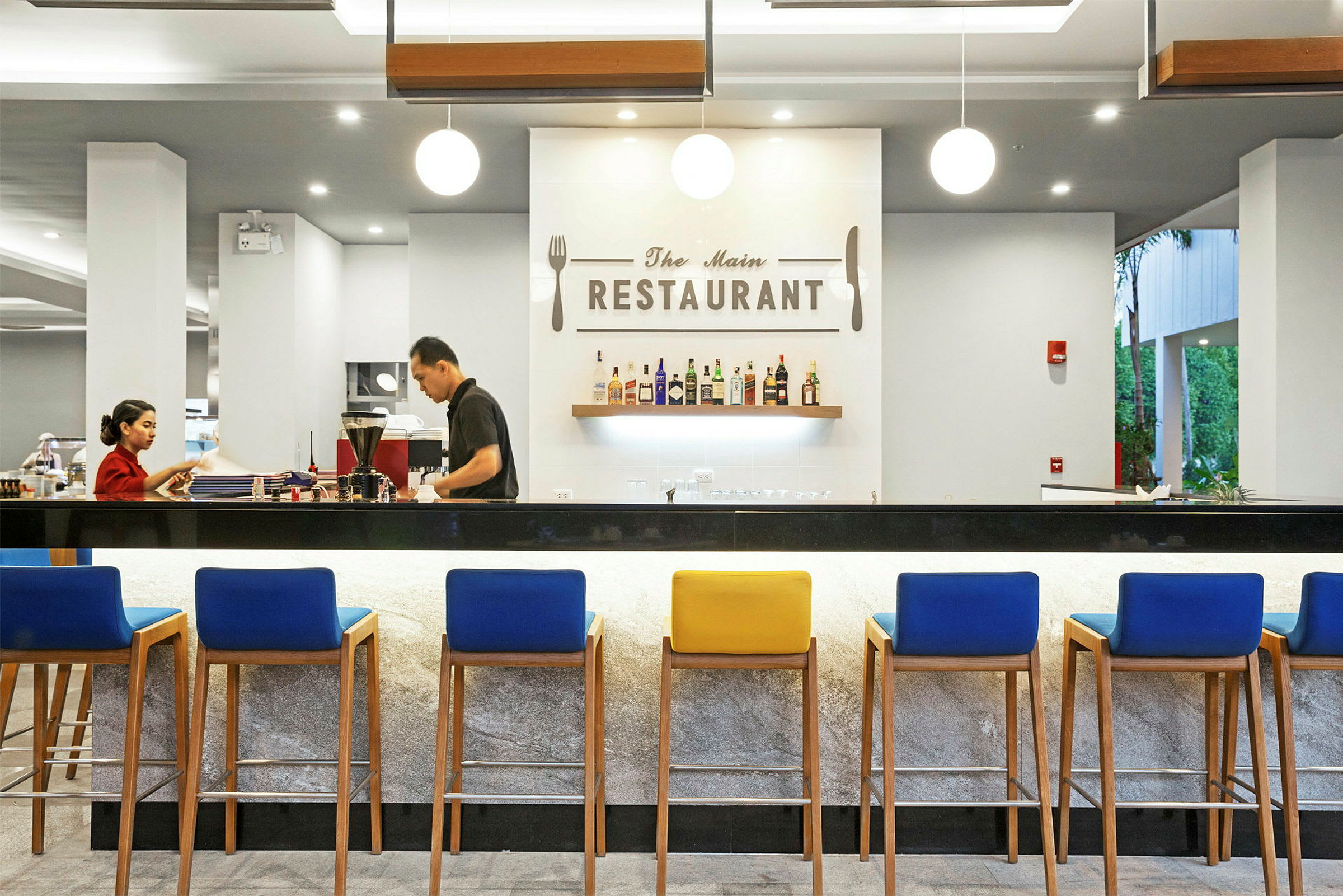 Tyylikäs ravintola modernilla sisustuksella, värikkäillä tuoleilla varustettu baari ja seinä, jossa on ripustettu pulloja sekä kyltti, jossa lukee 'The Main Restaurant'. (Tekoälyn luoma teksti)