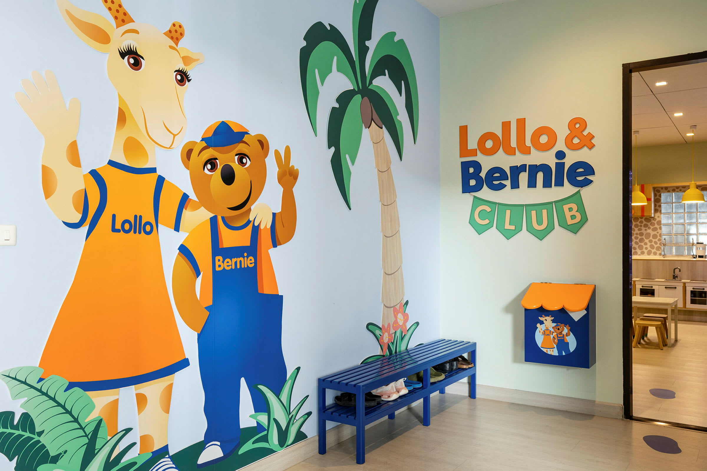 Värikäs seinämaalaus Lollo- ja Bernie-hahmoista seisoo seinällä lastenkerhossa. Huoneessa on myös sininen penkki ja kylttejä. (Tekoälyn luoma teksti)