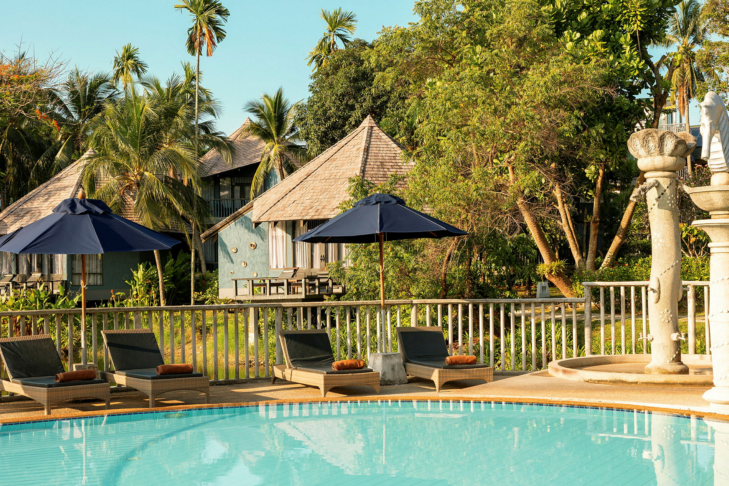 Uima-allas, jota ympäröivät aurinkotuolit ja varjot, taustalla puisia rakennuksia ja trooppisia puita. (Tekoälyn luoma teksti)