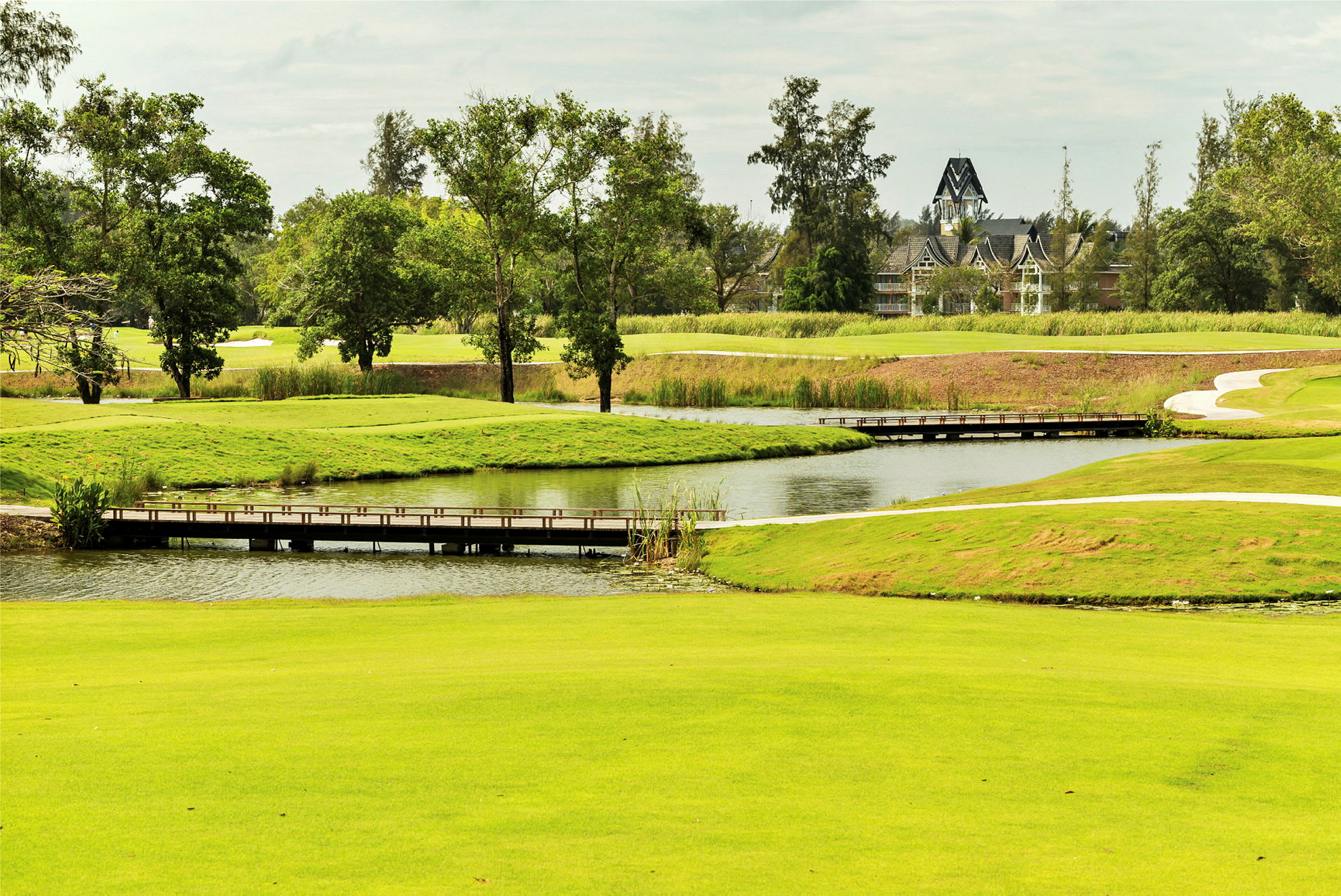 Kuva vihreästä golfkentästä, jossa on pieni kävelysilta, joka ylittää veden, ympäröitynä puilla ja kasvillisuudella. (Tekoälyn luoma teksti)