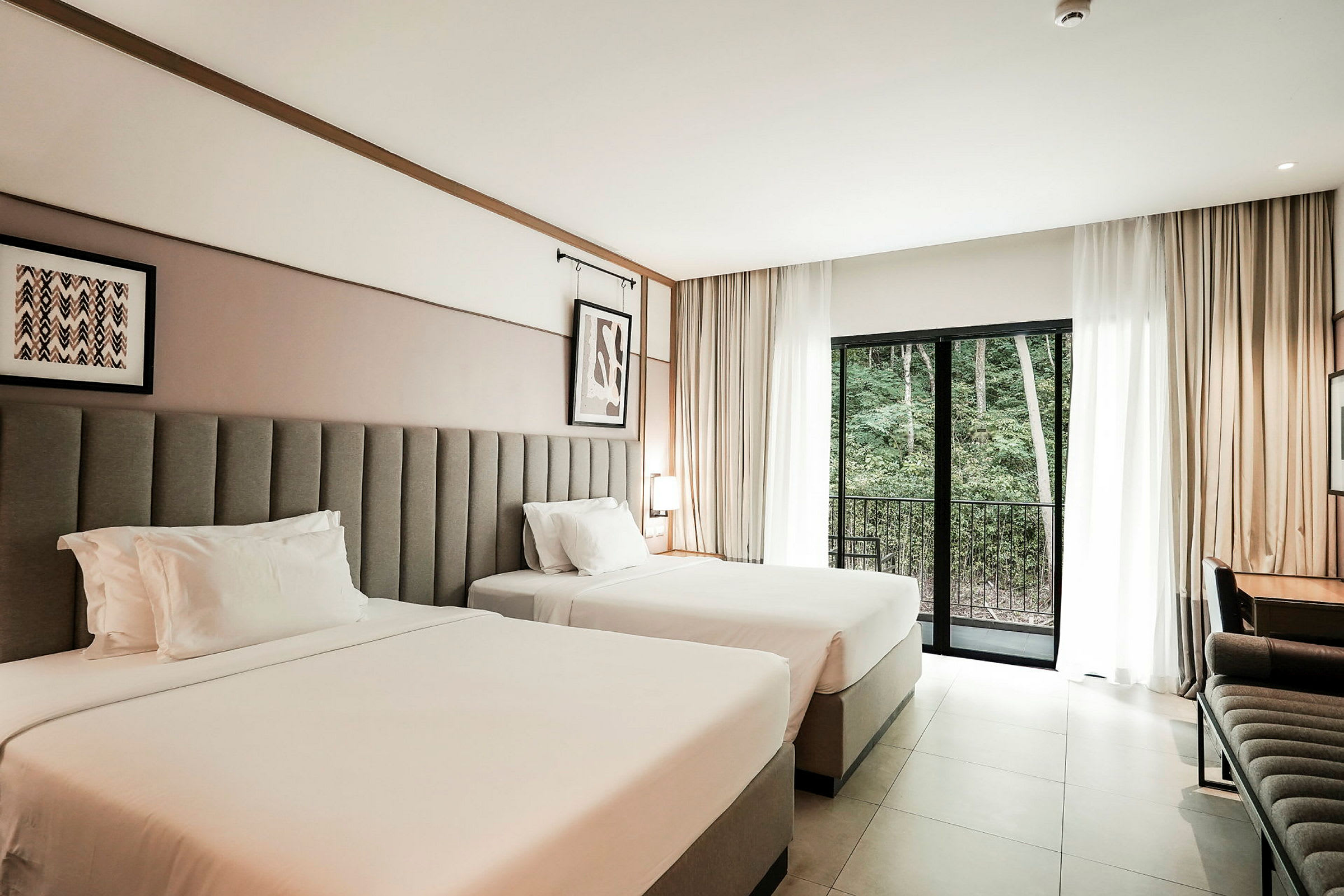 Hotellihuone, jossa on kaksi leveää sänkyä, kirjoituspöytä ja parvekkeen ovi näkymällä metsäalueelle. (Tekoälyn luoma teksti)