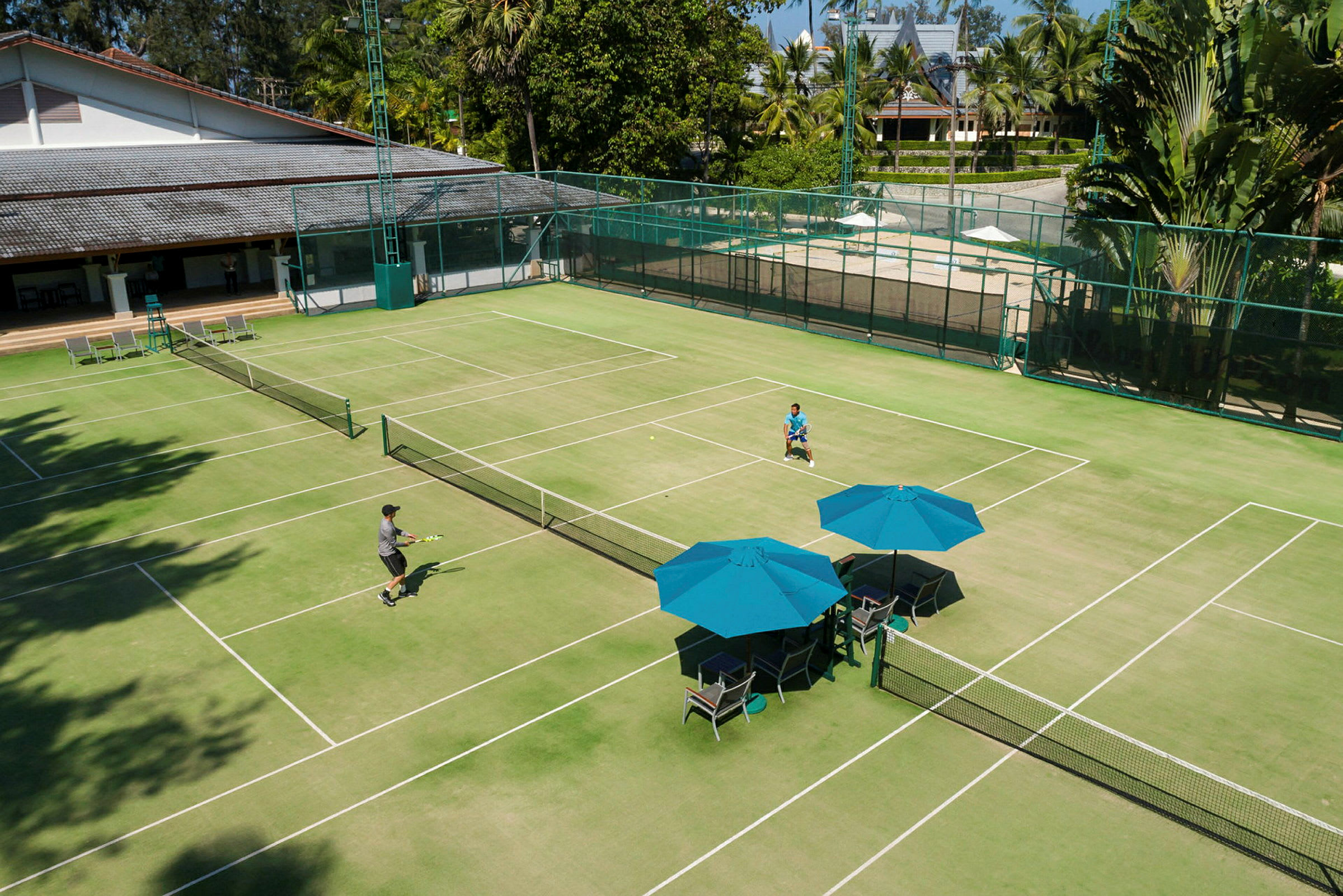 Vihreä tenniskenttä, jossa kaksi henkilöä pelaa tennistä. Kentän reunalla on sinisiä varjoja ja tuoleja. (Tekoälyn luoma teksti)