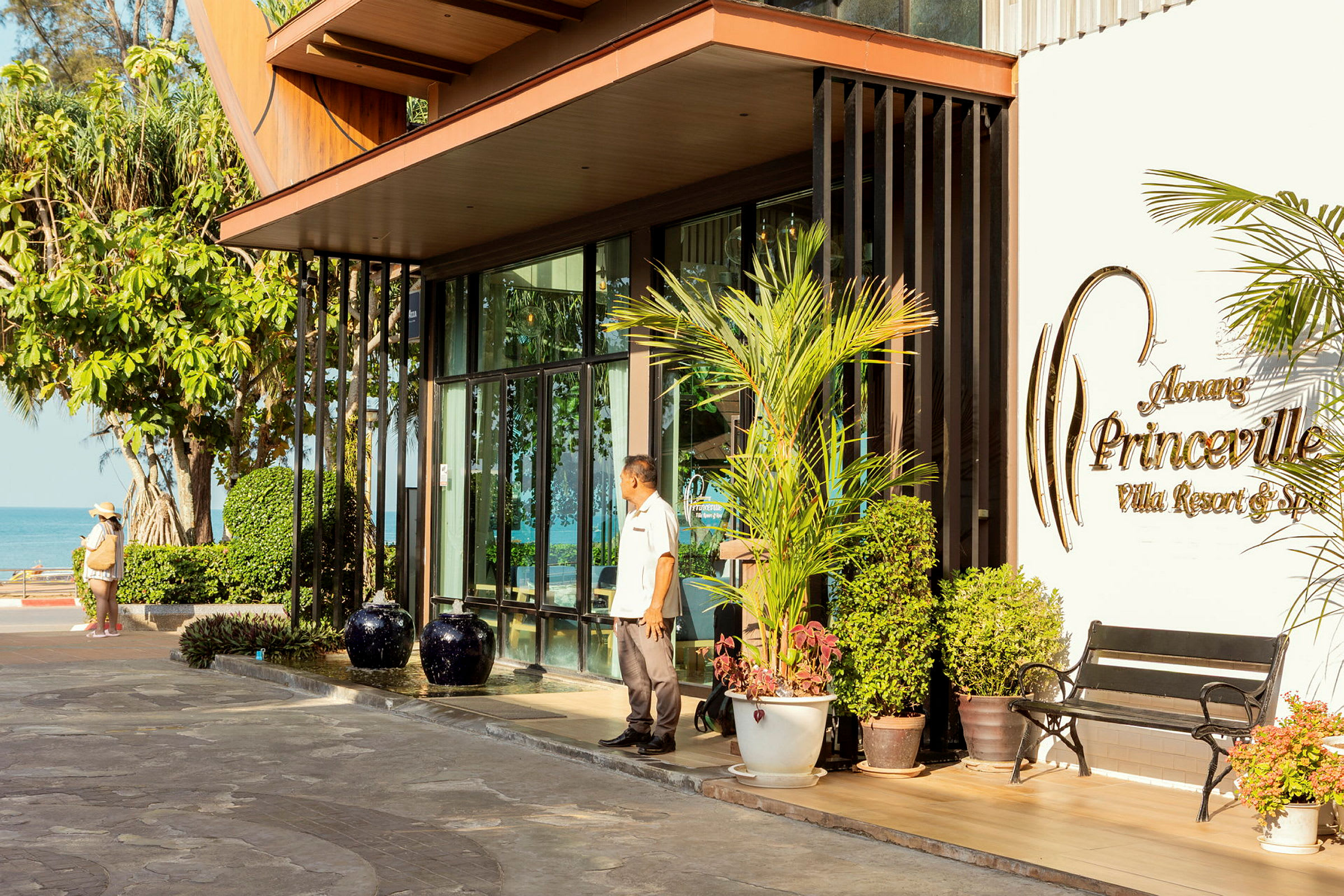 Lomakeskuksen sisäänkäynti, jossa on kyltti "Aonang Princeville Villa Resort & Spa". Ulkona näkyy istutuksia ja penkki, taustalla meri ja vehreys. (Tekoälyn luoma teksti)