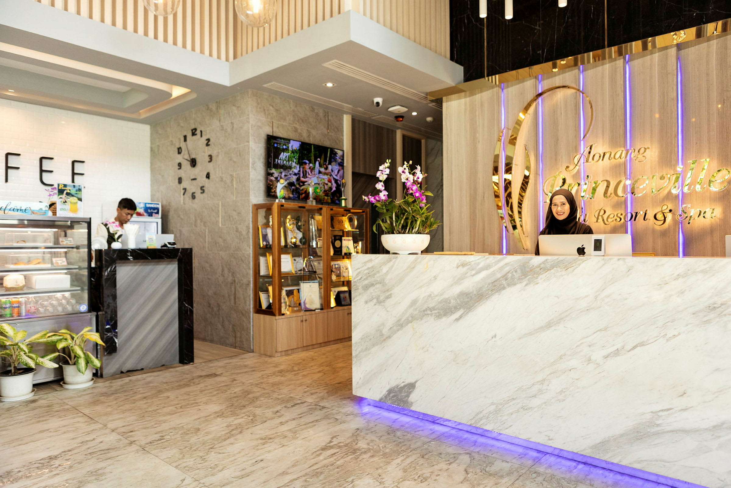 Vastaanotto, jossa on marmoritiski ja kyltti "Ao Nang Princeville Resort & Spa". Tiskin takana seisoo henkilö. Vasemmalla näkyy pieni kahvila ja vitriinikaappi palkintoineen. (Tekoälyn luoma teksti)
