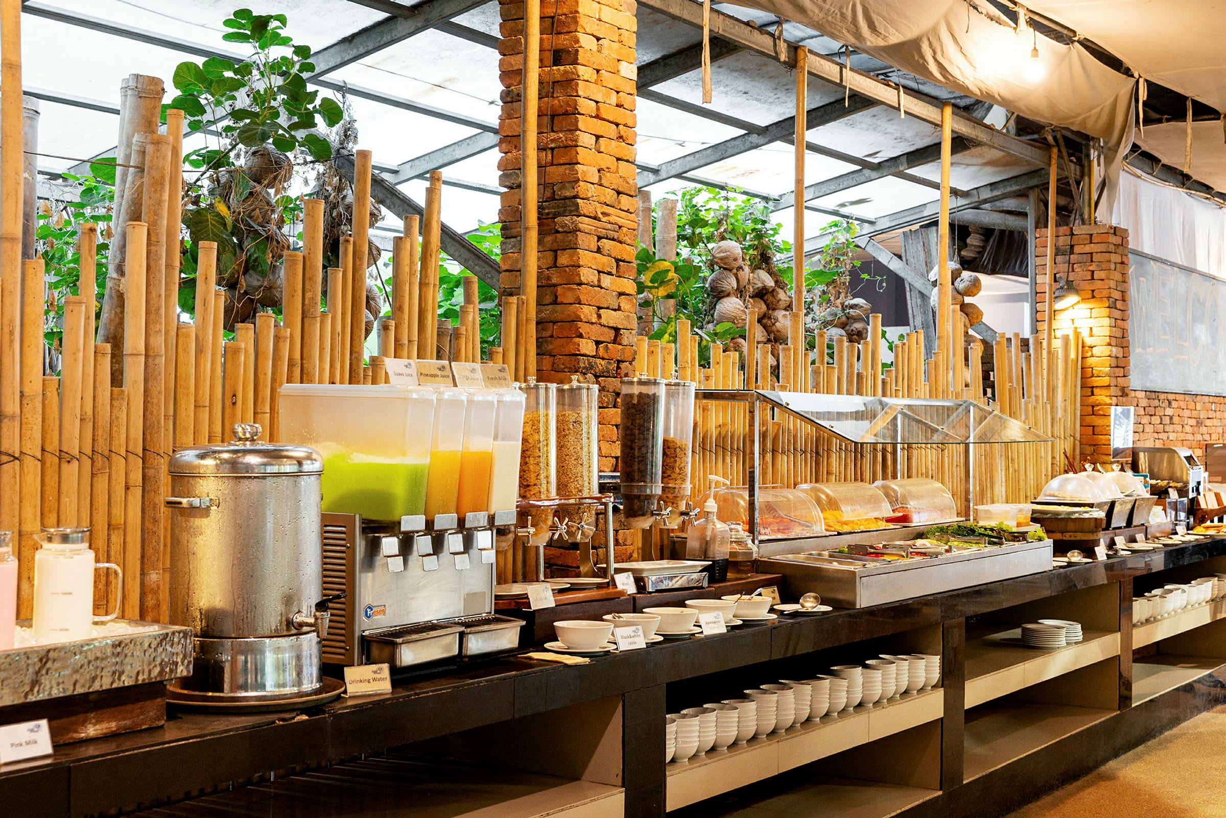 Buffet, jossa on erilaisia ruokavaihtoehtoja, kuten juomia, salaatteja ja aamiaisruokia, rustiikkisen sisustuksen edessä, jossa on bambu-yksityiskohtia ja eläviä kasveja. (Tekoälyn luoma teksti)
