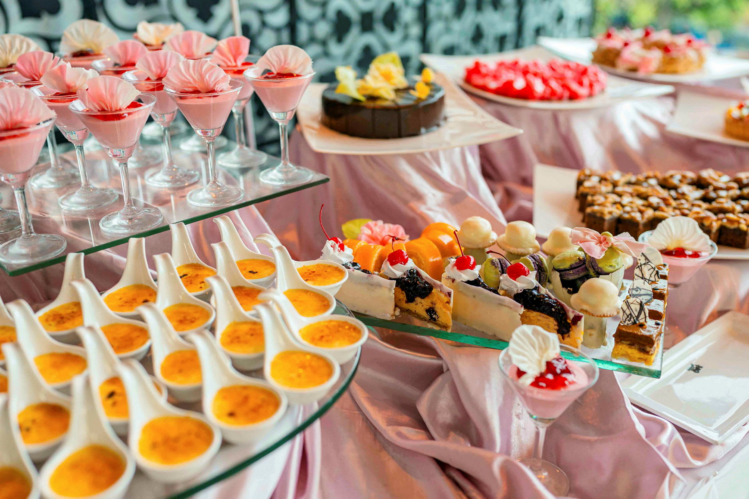 Buffet, jossa erilaisia jälkiruokia, kuten lasit vaaleanpunaisella mousseilla, crème brûléea ja koristeltuja leivonnaisia. (Tekoälyn luoma teksti)